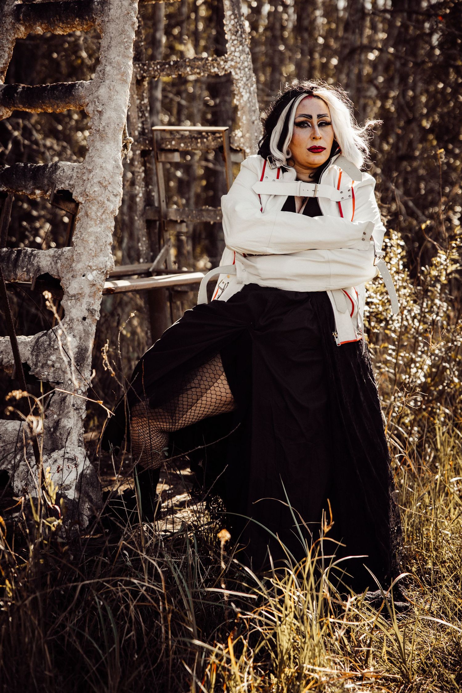 Woman in Cruella de Vil cosplay with black skirt and white jacket poses by rustic wooden structure.