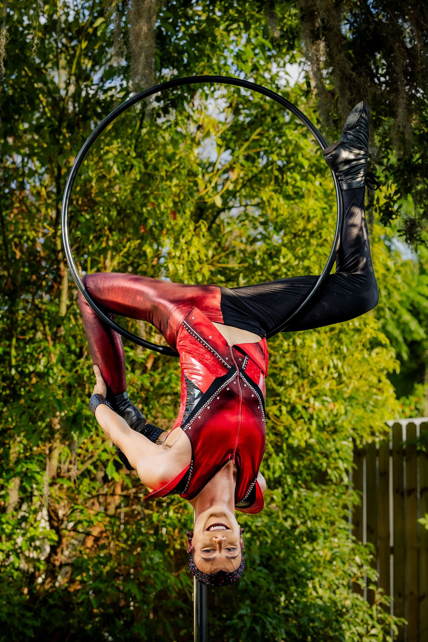 Aerial hoop performer in red costume balancing upside down on a lyra ring outdoors surrounded by green trees.
