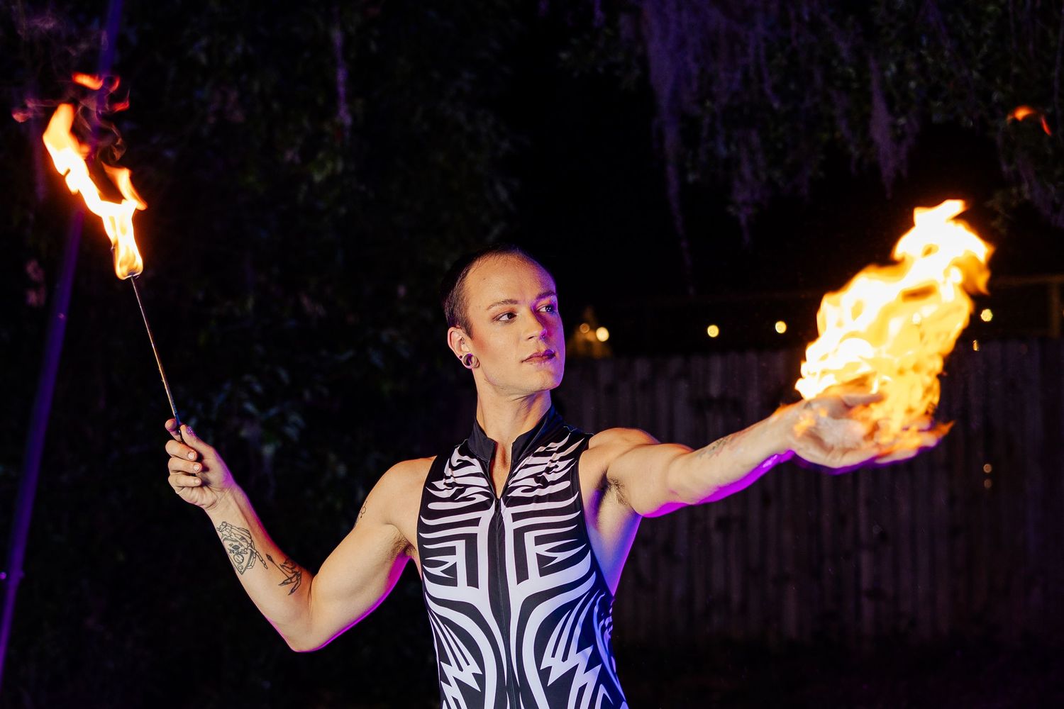 Fire performer in tribal print bodysuit holds two flaming torches during an outdoor nighttime performance.