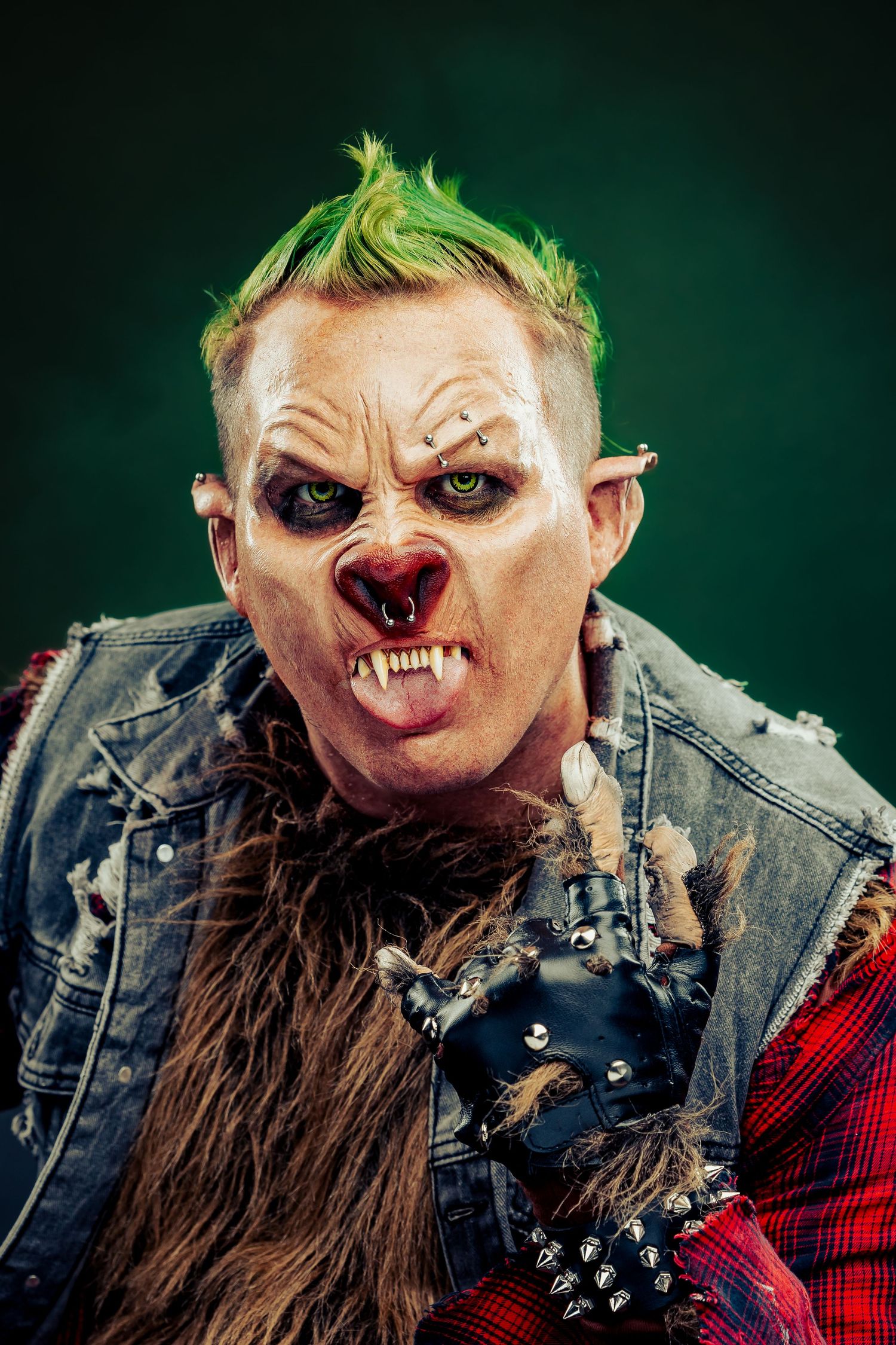 Scary punk troll Halloween costume with green mohawk, prosthetic makeup, fangs, and spiked leather gloves on dark background.