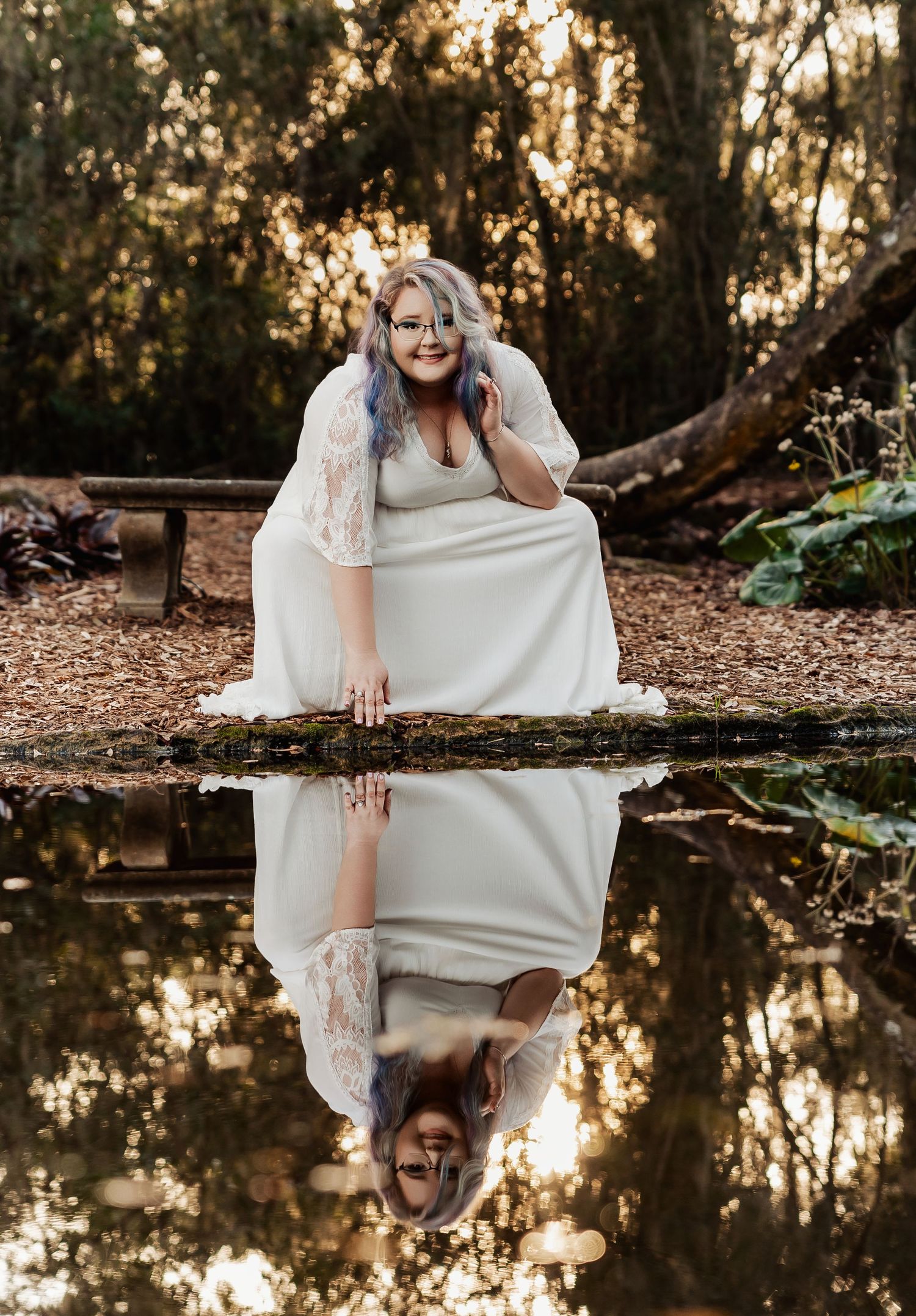 Plus-size bride in white lace gown with purple hair poses by reflective pond in woodland setting.