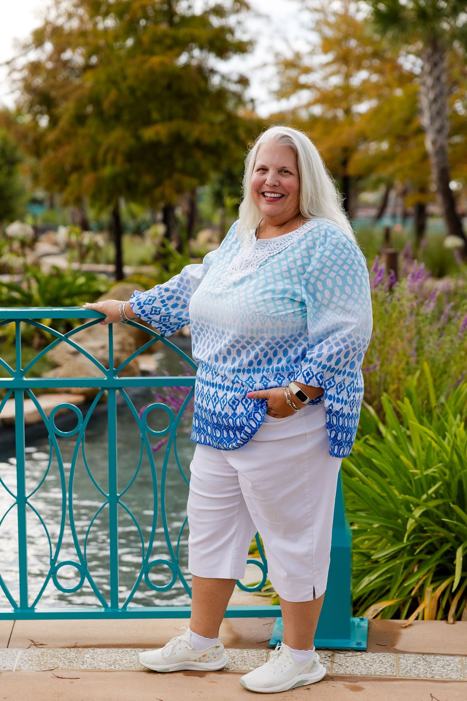 Smiling woman in blue patterned sweater and white shorts poses on teal bridge in lush green park.