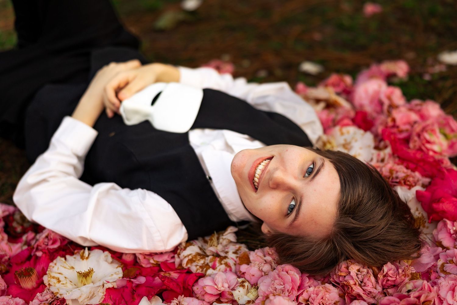 Smiling young woman lying among pink and red flowers wearing a black vest and white shirt in a garden setting.