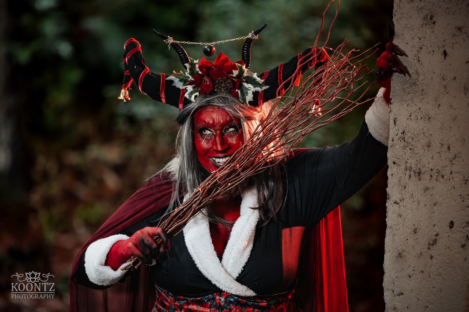 A cosplayer with red face paint, antler crown, and red cape holds sparkling branches in a dark forest setting.