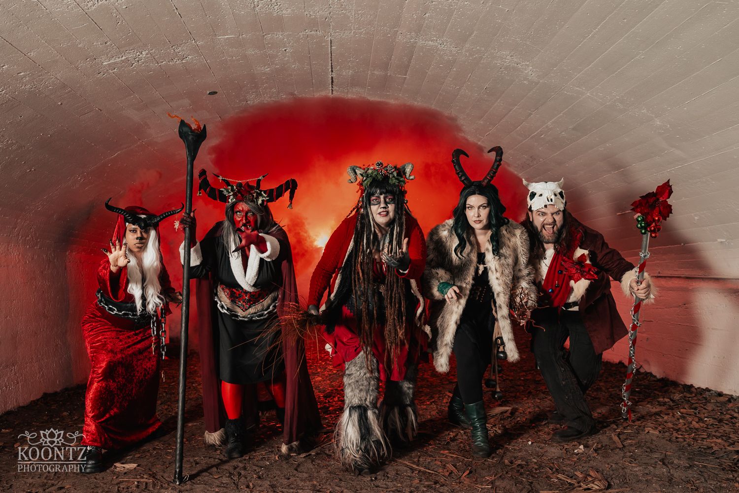 Five people in elaborate dark fantasy devil costumes pose in a tunnel with dramatic red lighting.