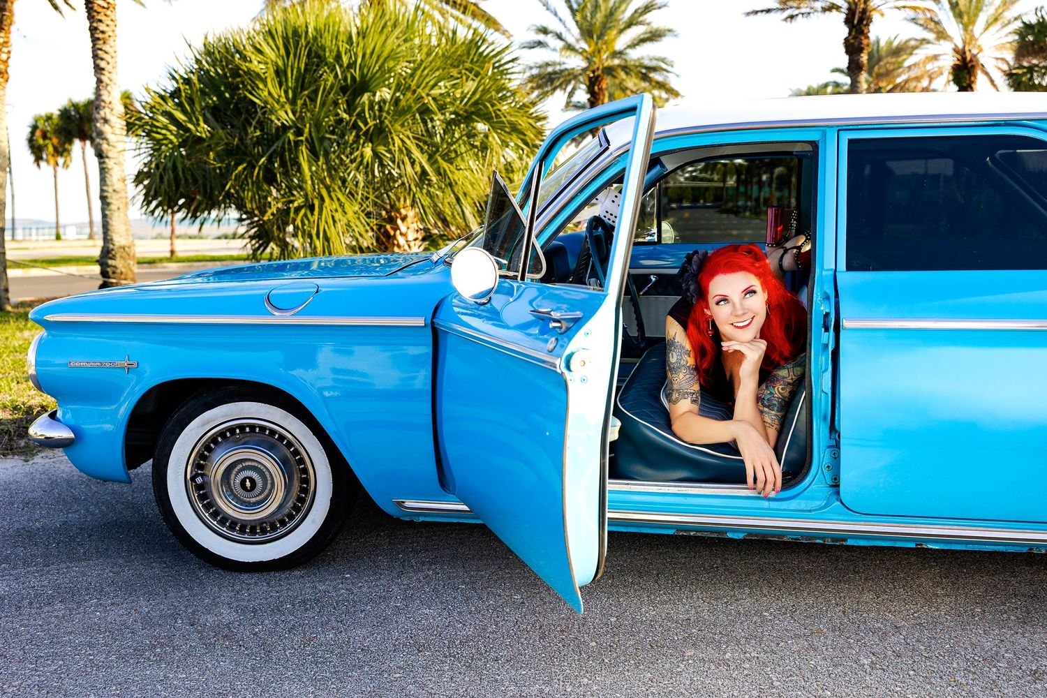 Smiling tattooed redhead woman leans out window of classic blue vintage car surrounded by palm trees.