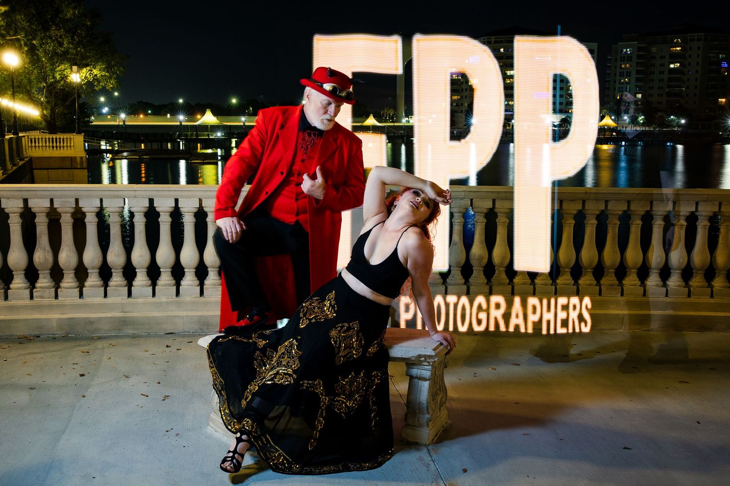 Two people pose at night beside large EPP Photographers sign near a waterfront railing.