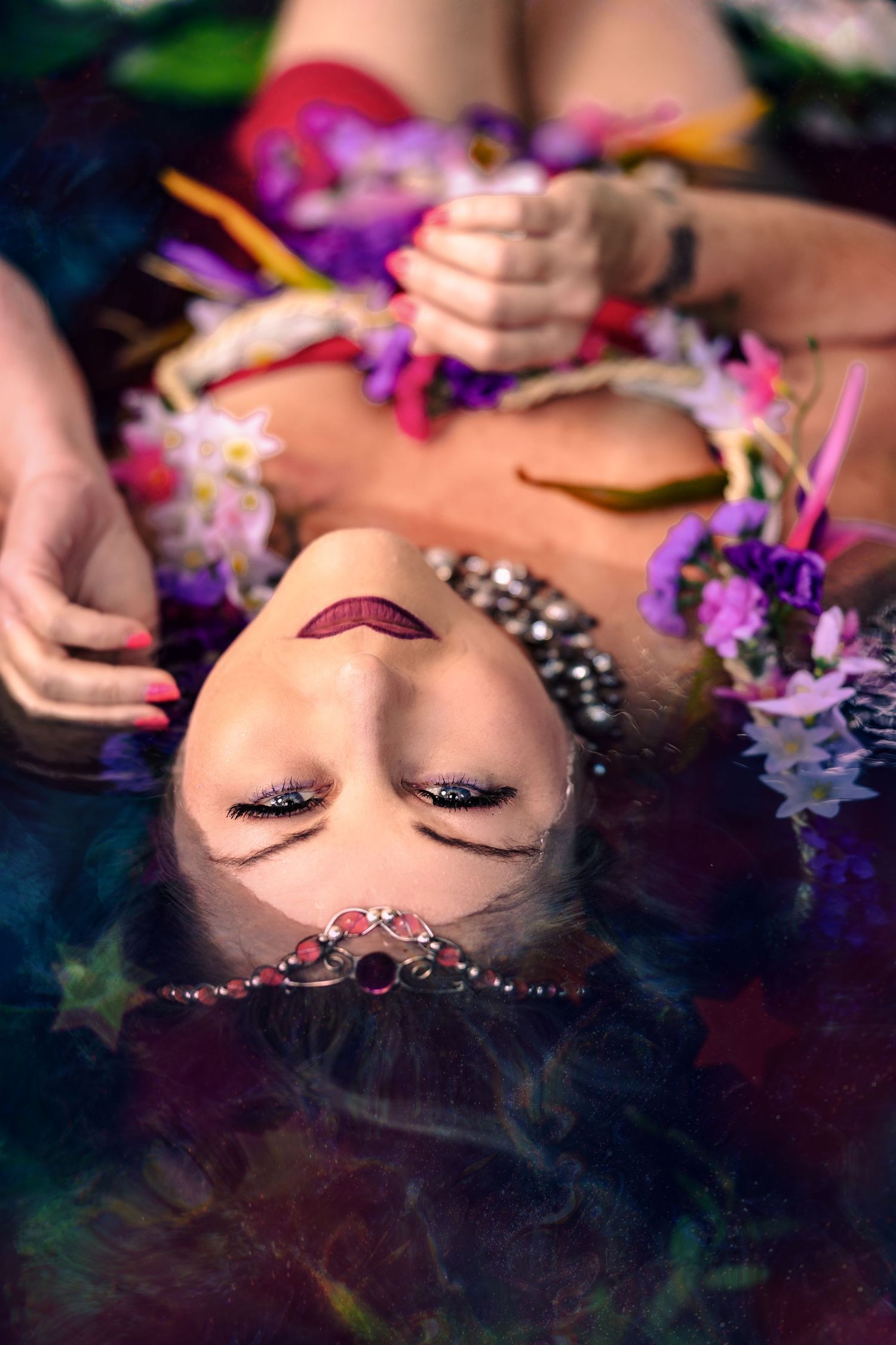 Woman with dark lipstick and jeweled nose piece lies in water surrounded by colorful flowers.