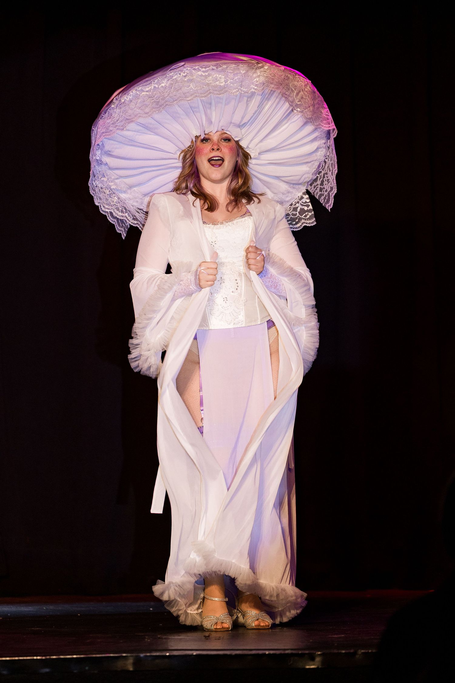 A smiling performer in an elaborate white gown and large hat stands on a dark stage.