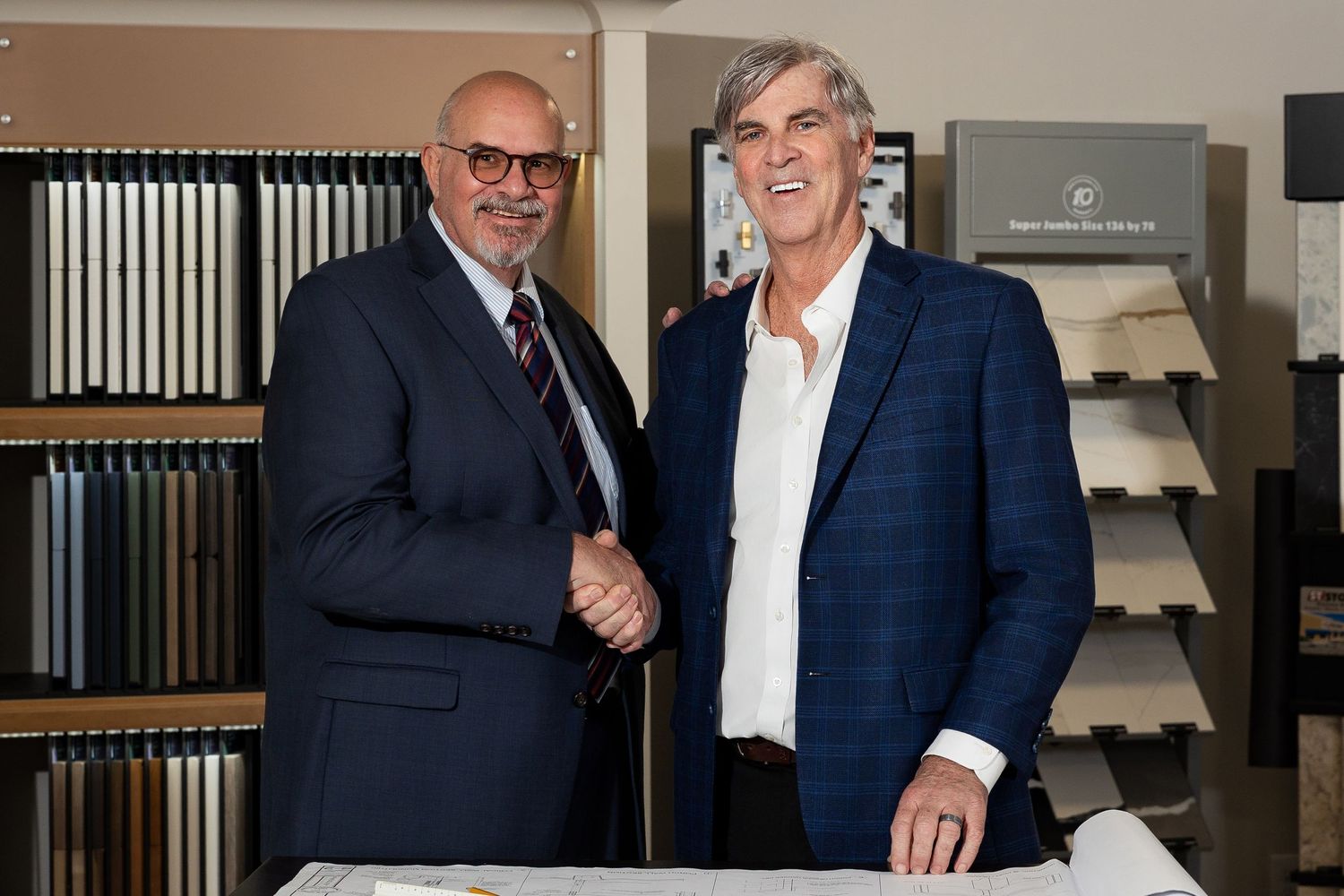 Two businessmen shaking hands in a tile showroom with material samples in the background.