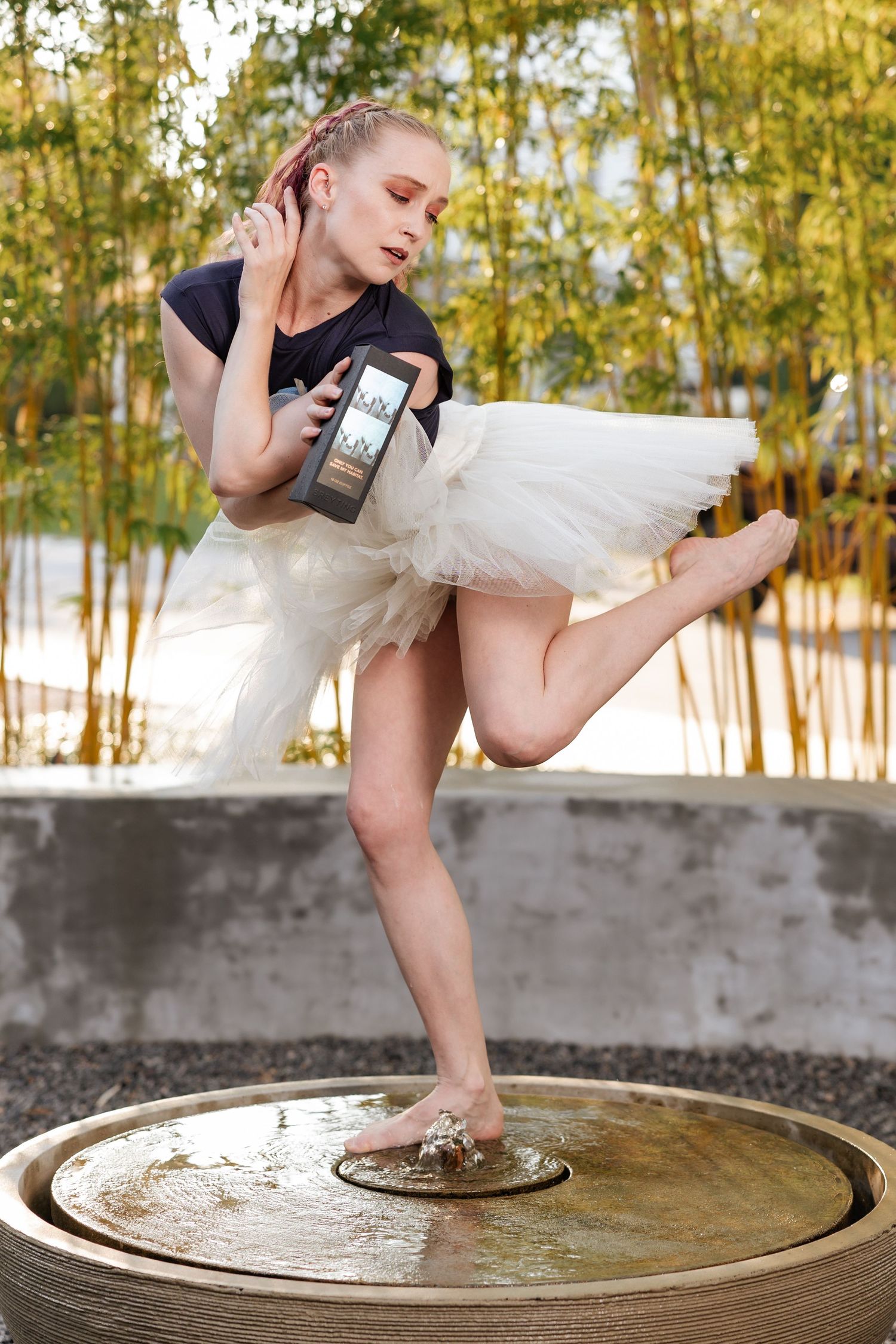 Ballerina in white tutu balancing on a water fountain bowl while holding a smartphone outdoors.