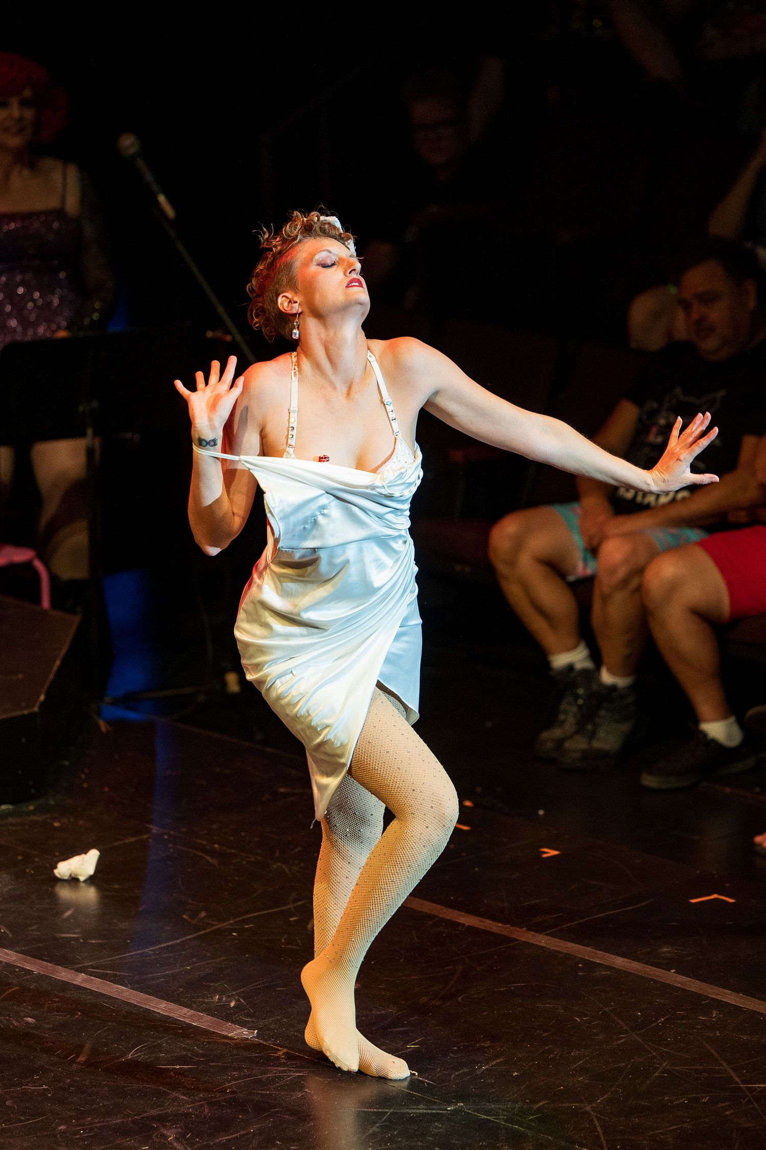 Barefoot burlesque dancer in white slip dress performs expressive routine before seated cabaret audience.