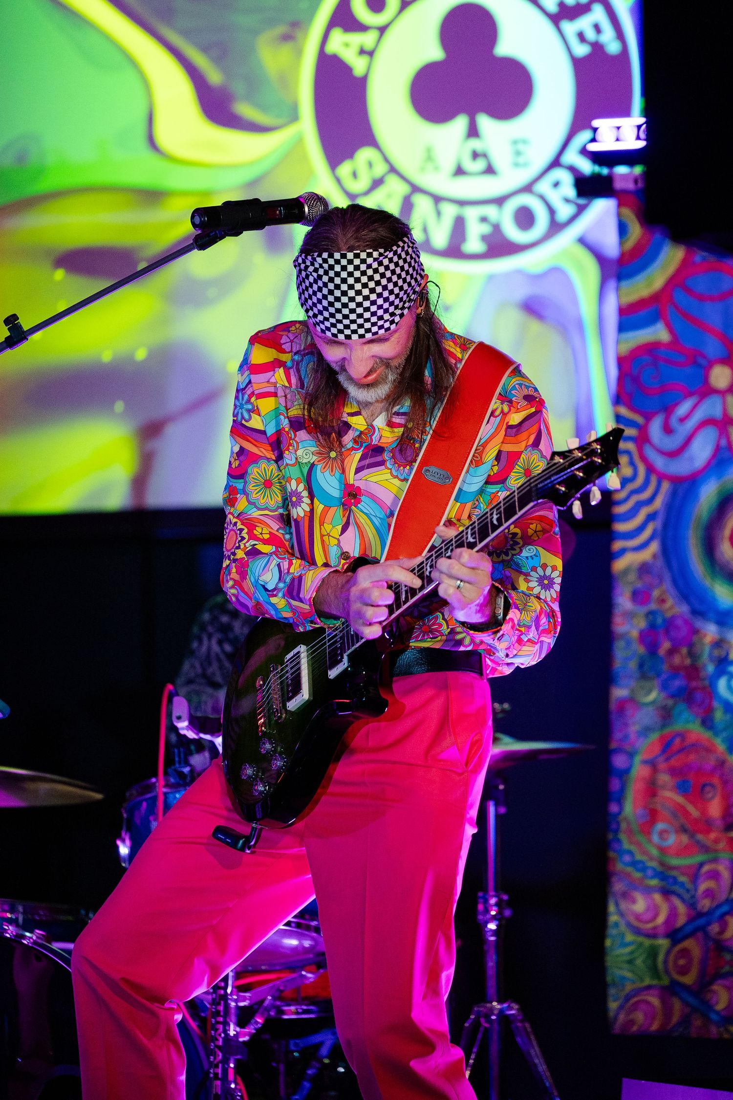 Guitarist in colorful psychedelic outfit and checkered headband performing on stage with red guitar.