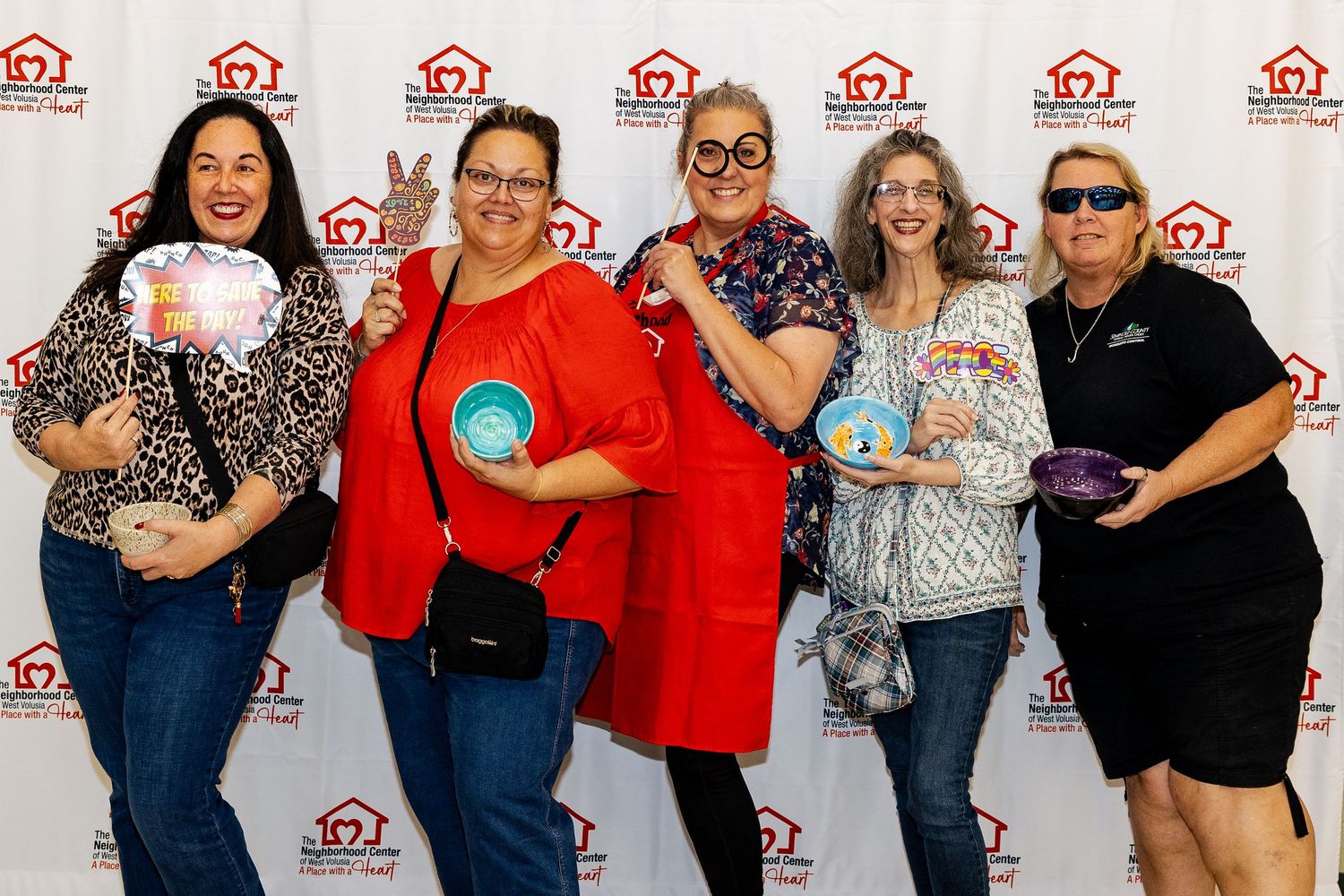 Five women pose playfully with props in front of a Neighborhood Housing Fund step-and-repeat banner.