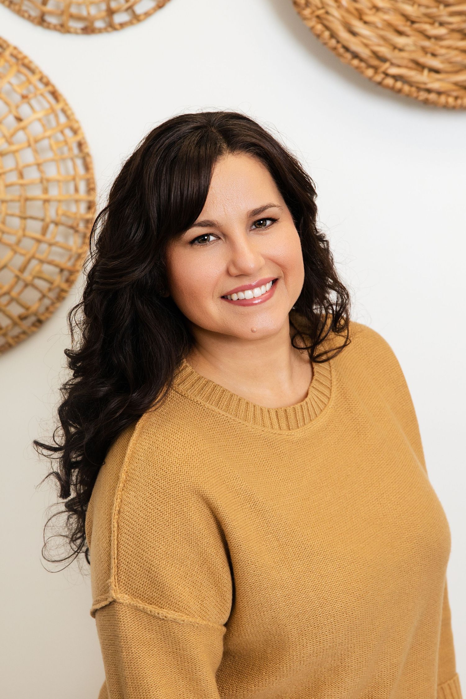 Young brunette woman with curly hair in mustard yellow sweater smiling against white wall decor.