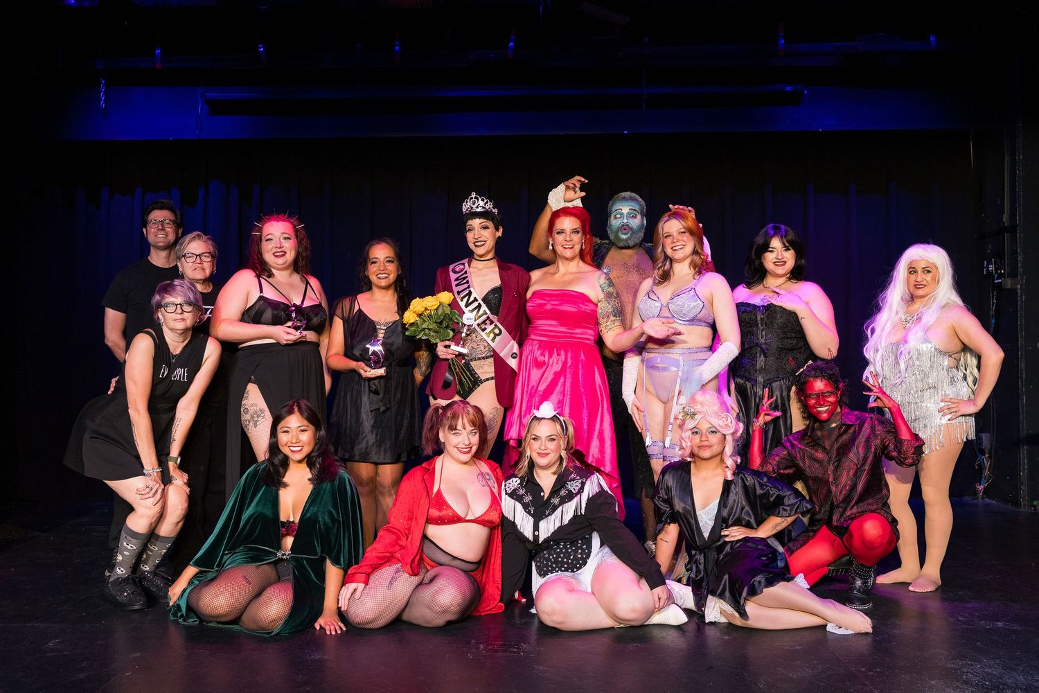 Group photo of burlesque performers on stage, with crowned winner in pink gown holding bouquet at center.