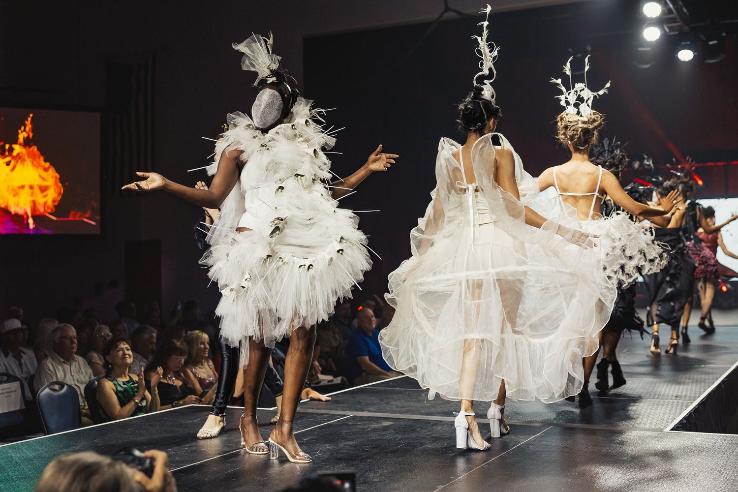 Models in elaborate white feathered fantasy gowns walk runway at creative fashion show event.