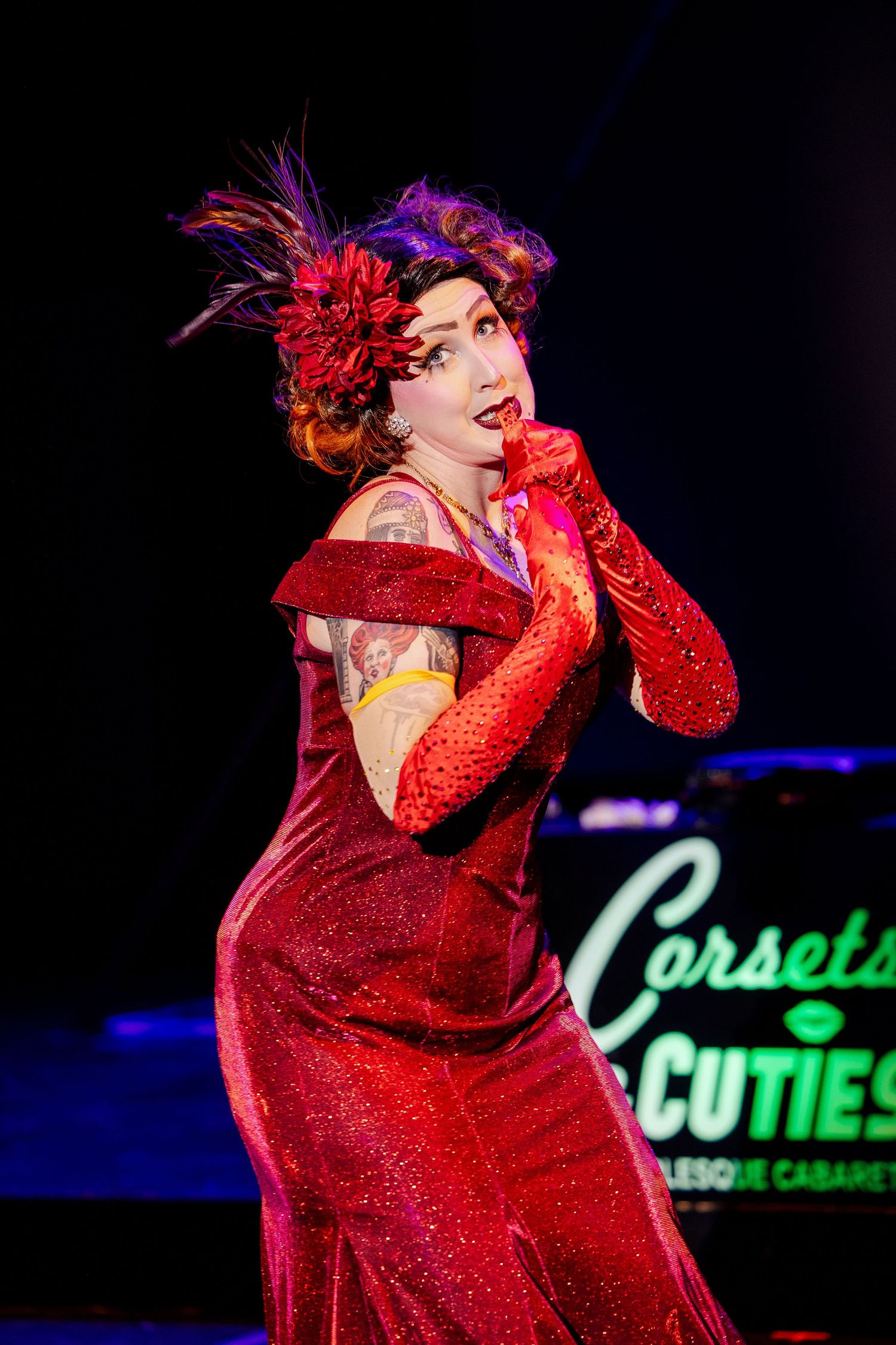 A burlesque performer in a red glittering gown with feathered headpiece poses on stage at Corsets and Cuties Cabaret.