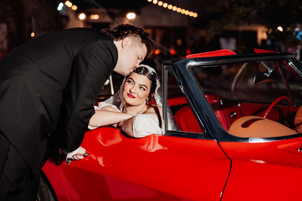 Groom kissing bride in a red convertible car at night during a romantic wedding photo shoot.