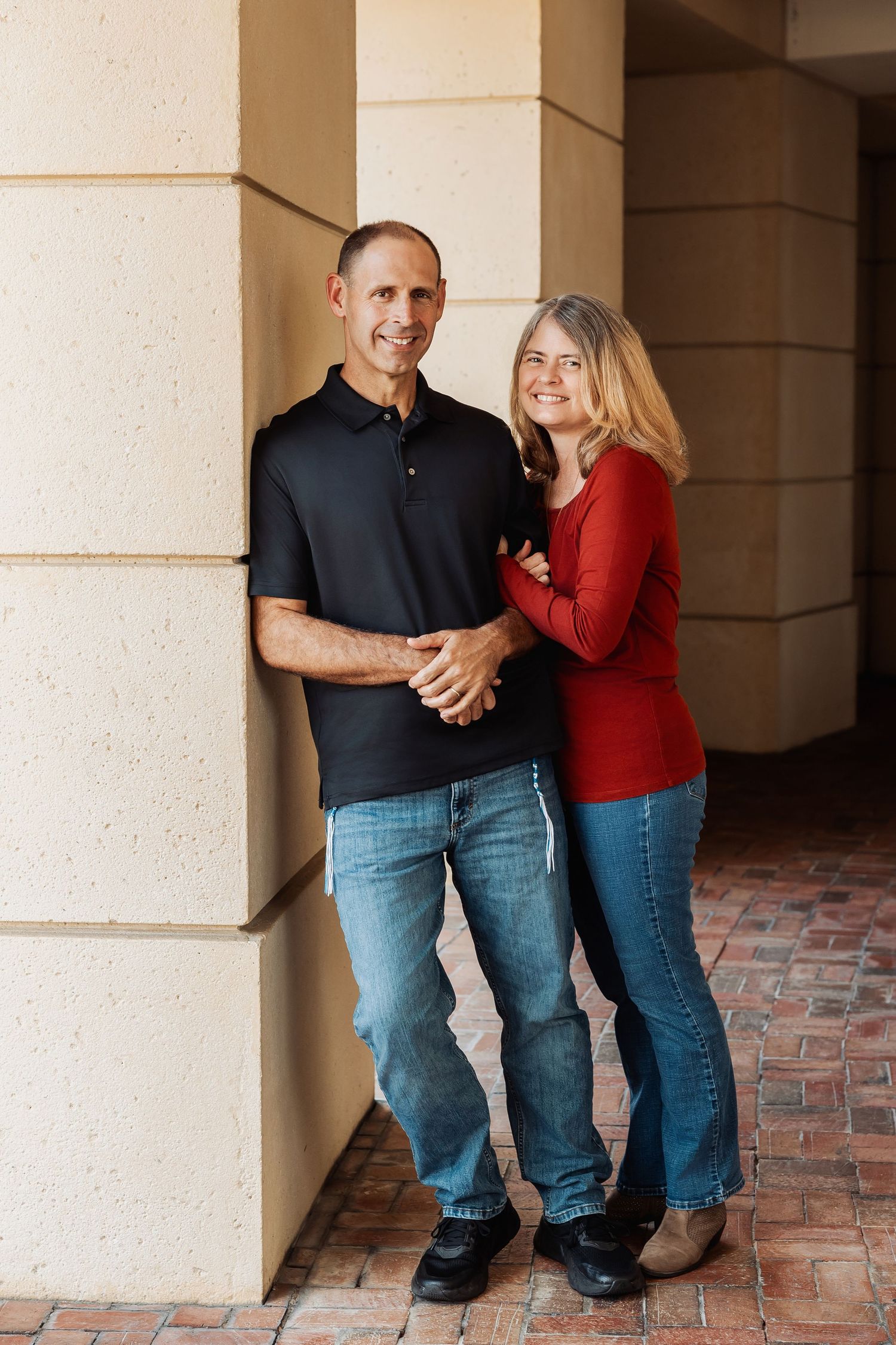 Smiling couple posing together near stone columns, man in black polo and woman in red top.