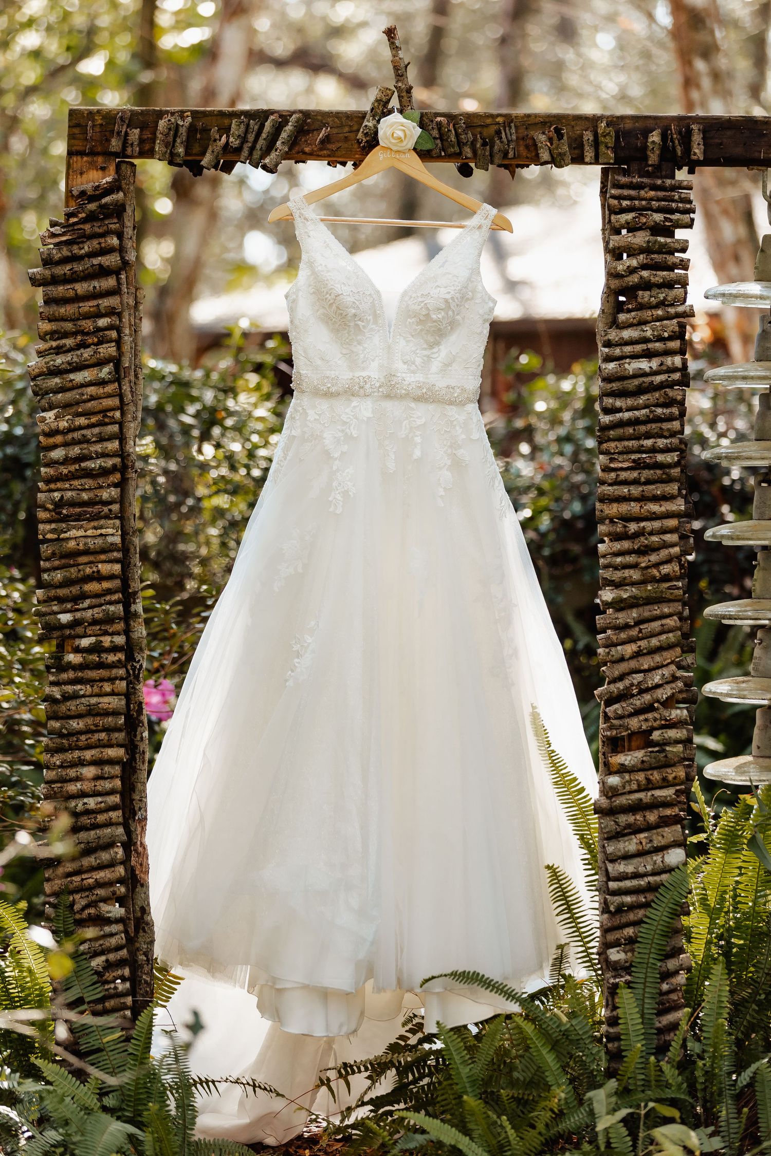 White lace wedding dress hanging on rustic wooden frame in garden setting with ferns and natural lighting.