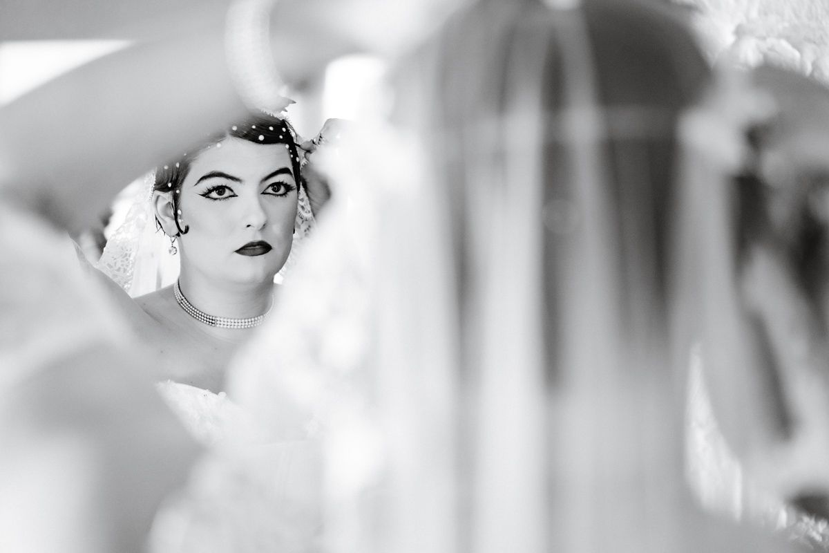 Black and white close-up of bride with dramatic makeup reflected in dressing room mirror.