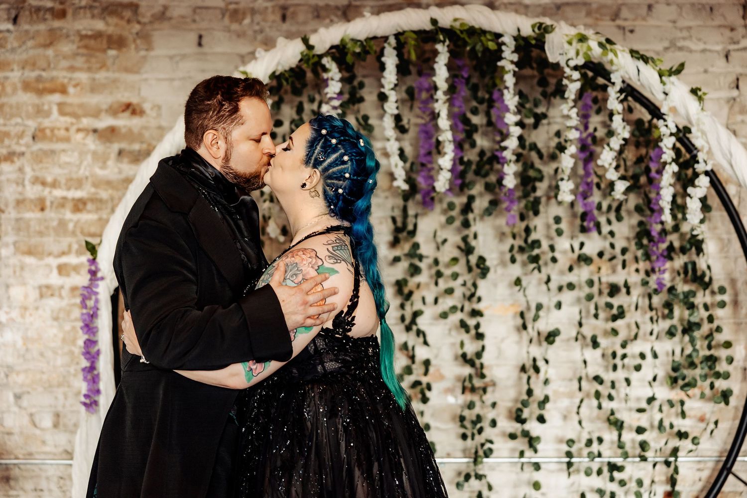 Couple sharing first kiss in black wedding attire beneath floral circular arch with greenery.