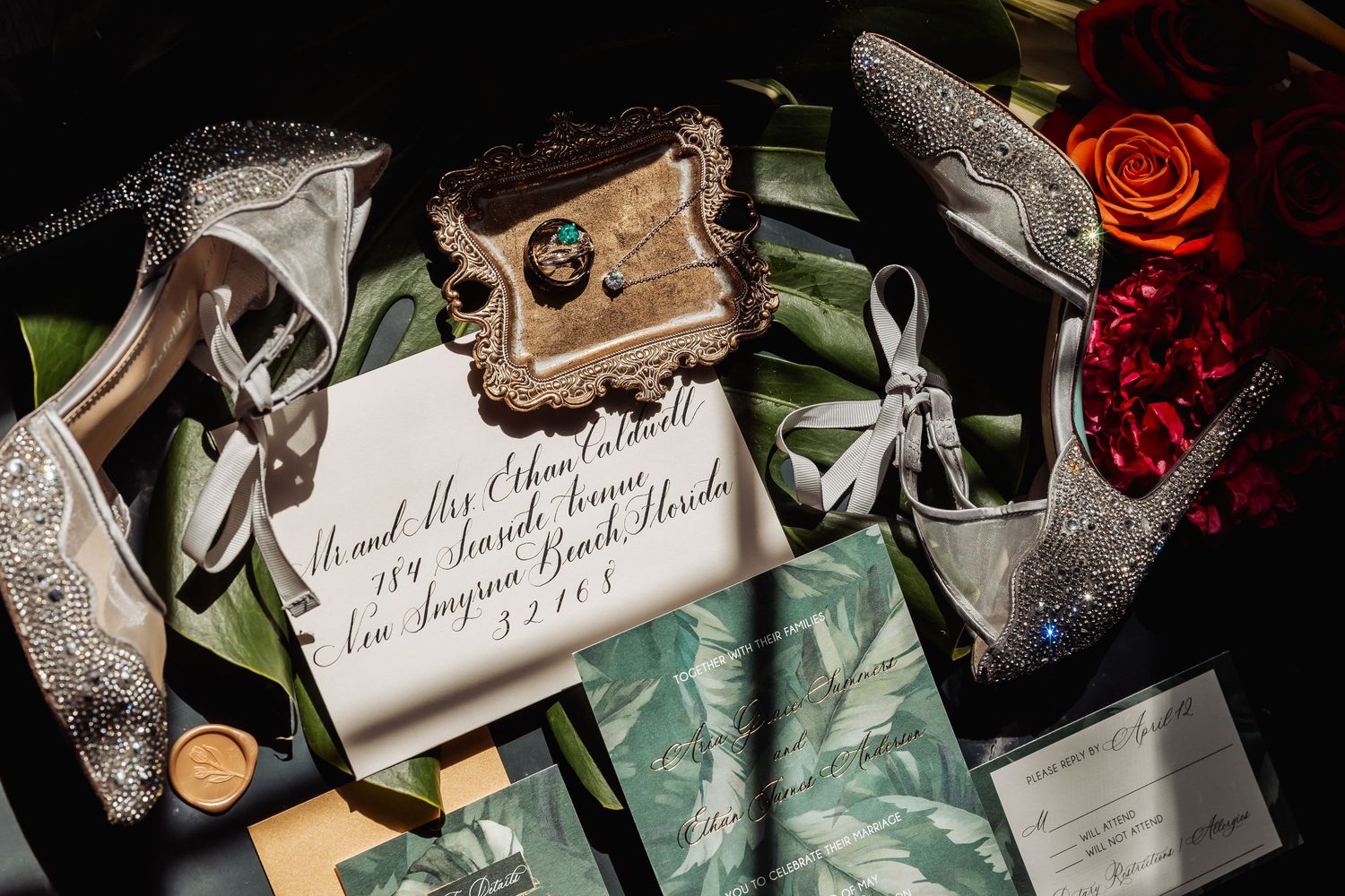 Elegant wedding flat lay with glittery heels, invitation, rings, and red roses on dark surface.