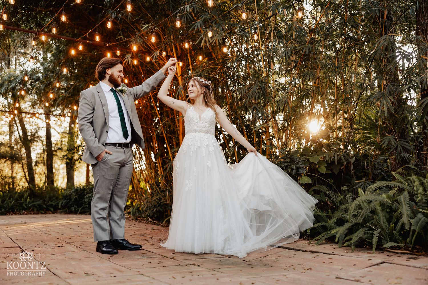 Bride and groom dance under glowing string lights at golden hour in a lush outdoor garden wedding setting.
