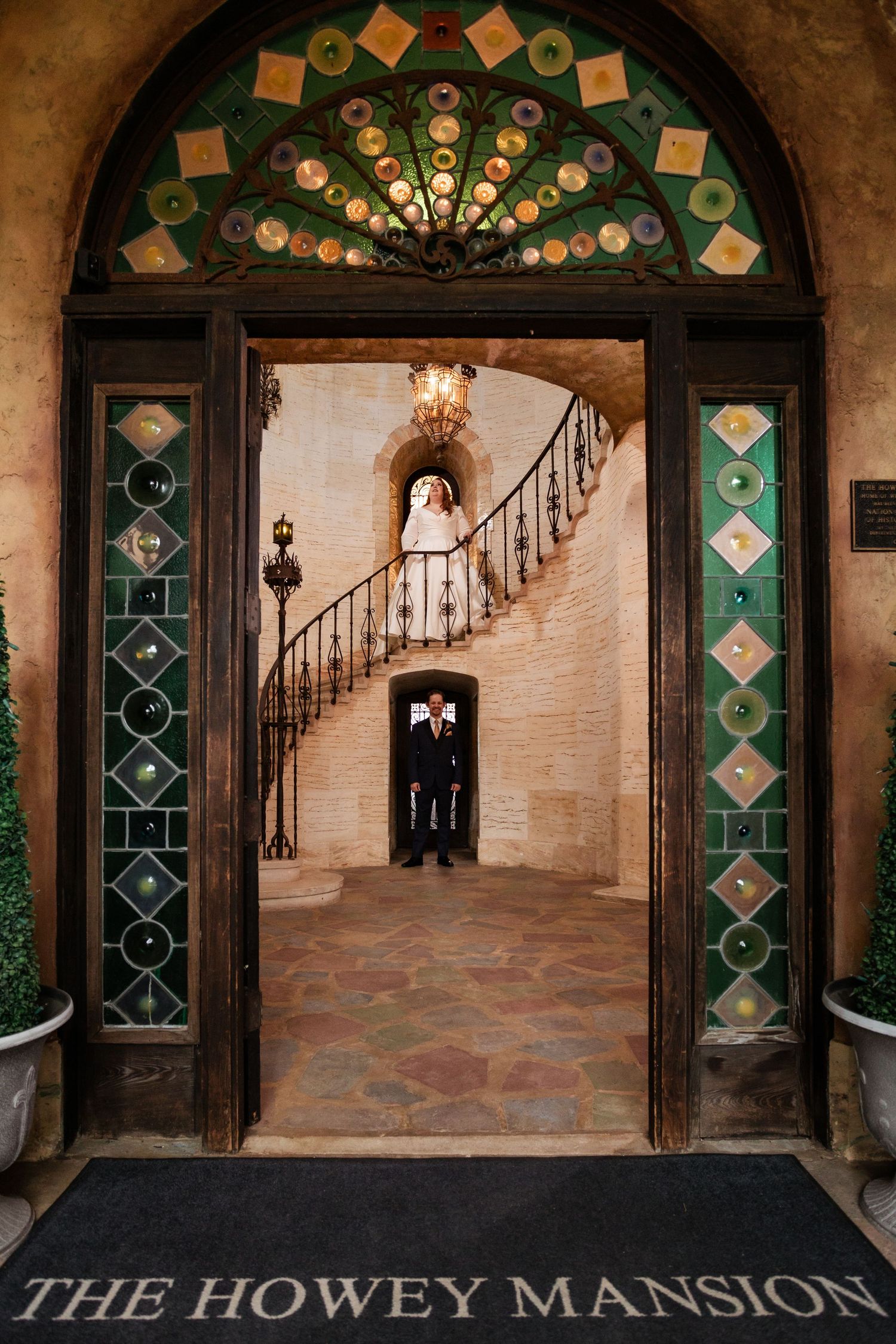 Bride and groom posing on a grand spiral staircase viewed through the ornate doors of The Howey Mansion.