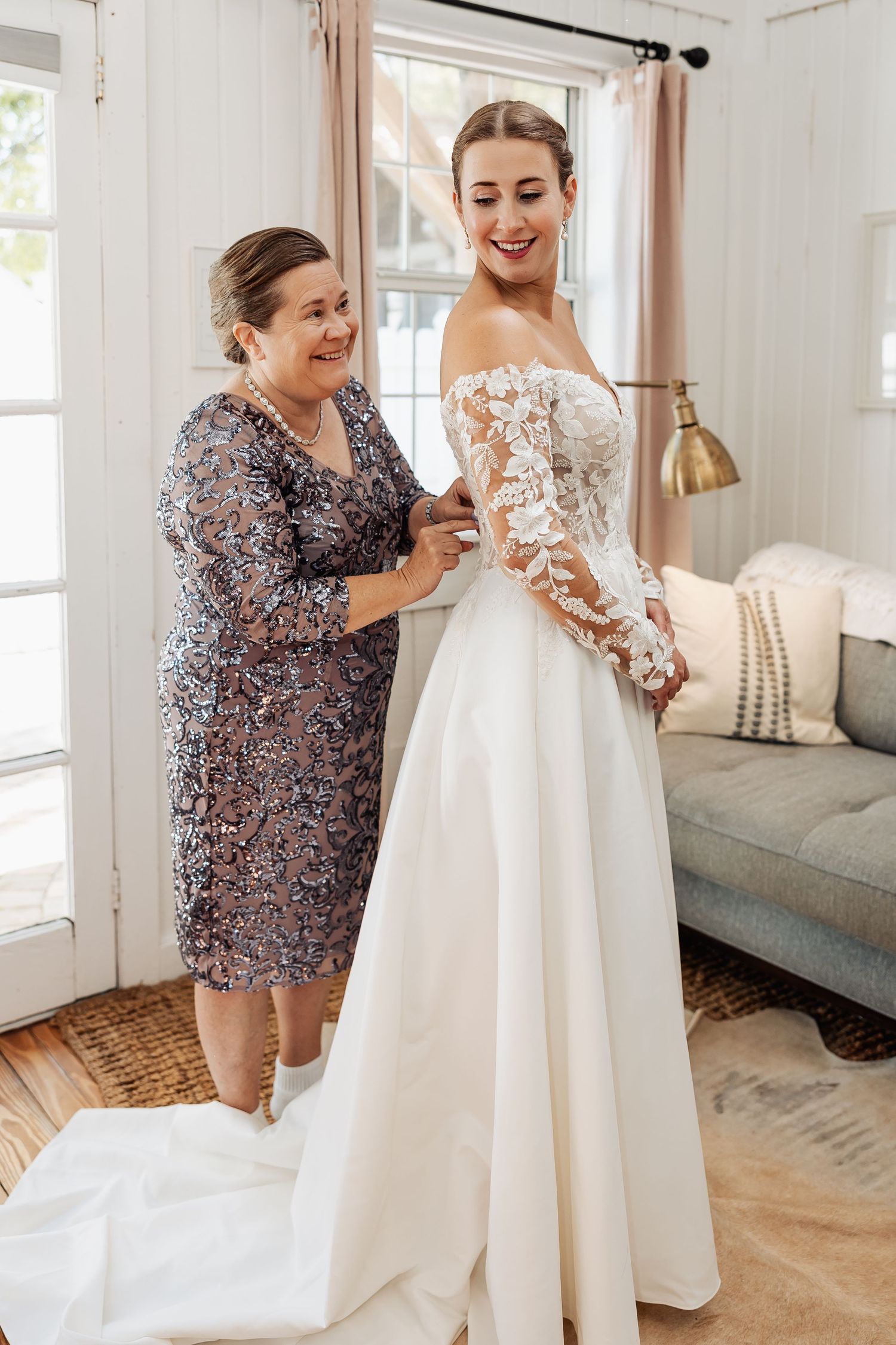 Mother helping bride button her long-sleeve lace and satin wedding gown in a bright white room.