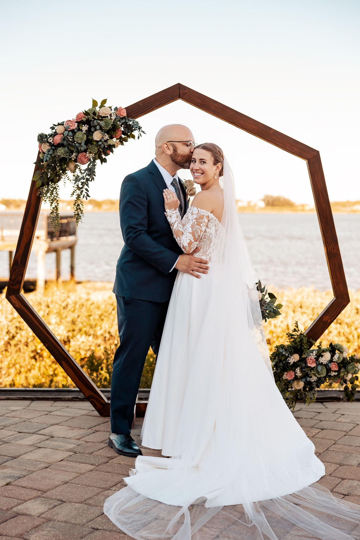 Bride and groom posing before hexagonal wooden arch with floral accents near waterfront.