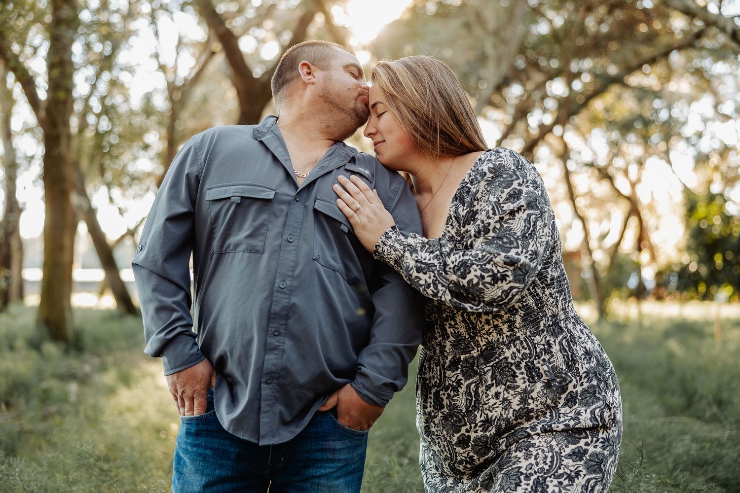 An engaged couple shares a tender forehead kiss moment surrounded by trees in golden light.