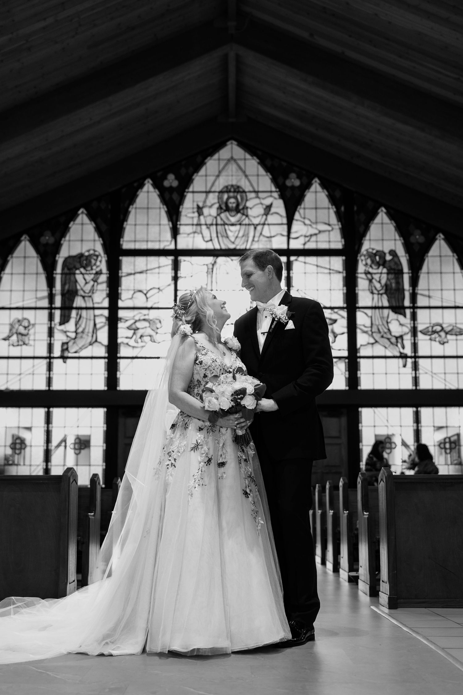 Black and white portrait of bride and groom embracing before stunning stained glass church windows.