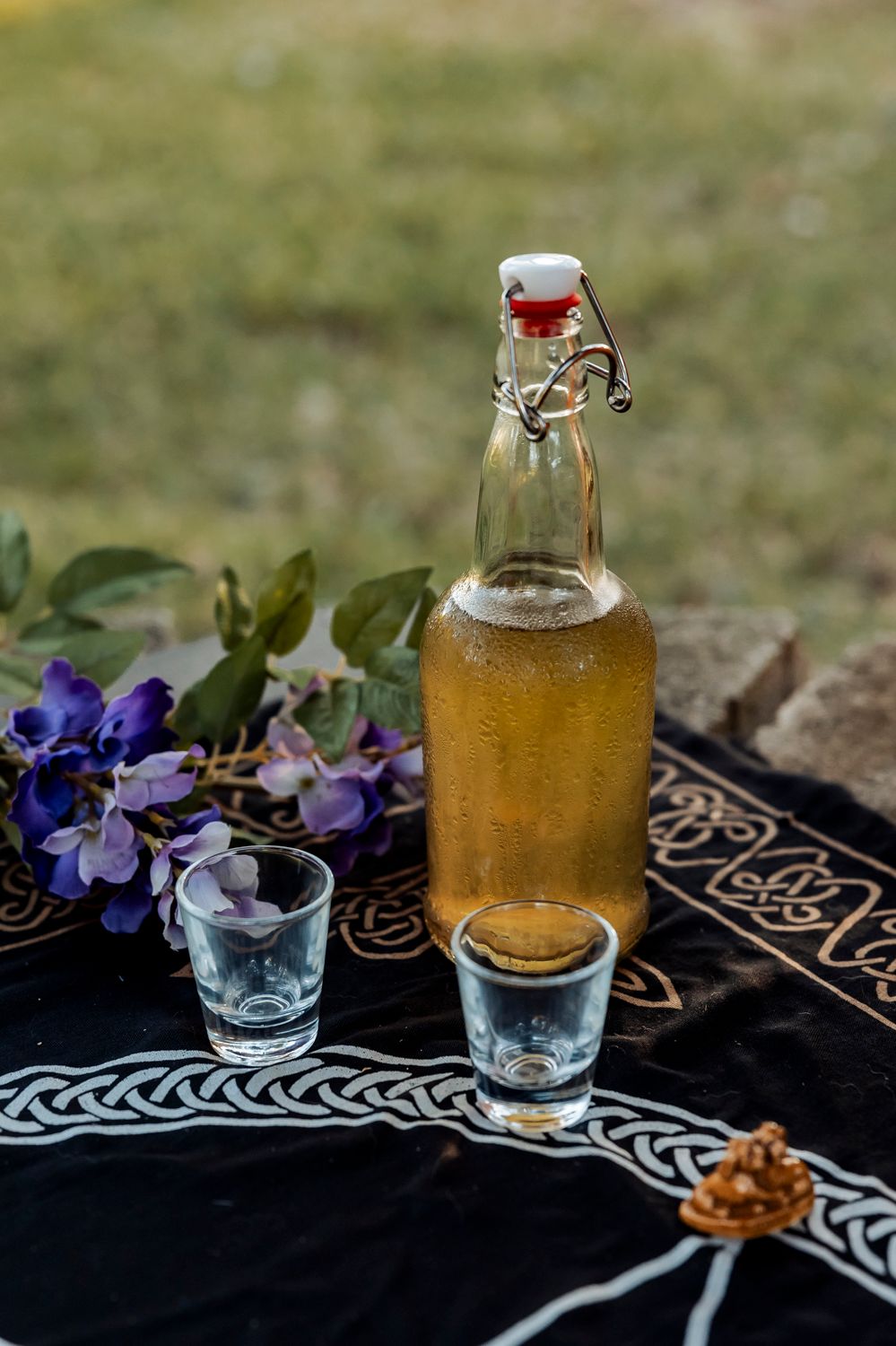 Amber liquid in vintage bottle with shot glasses on decorated cloth with purple flowers.
