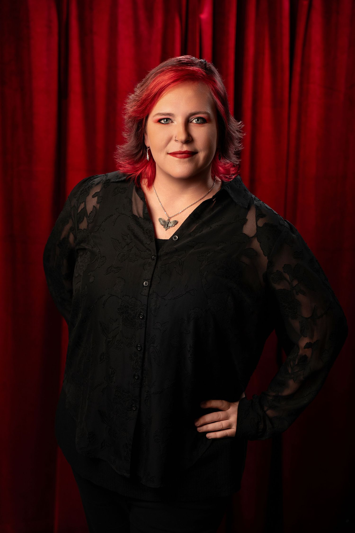 Woman with red and gray hair wearing black lace blouse posing against a red curtain backdrop.