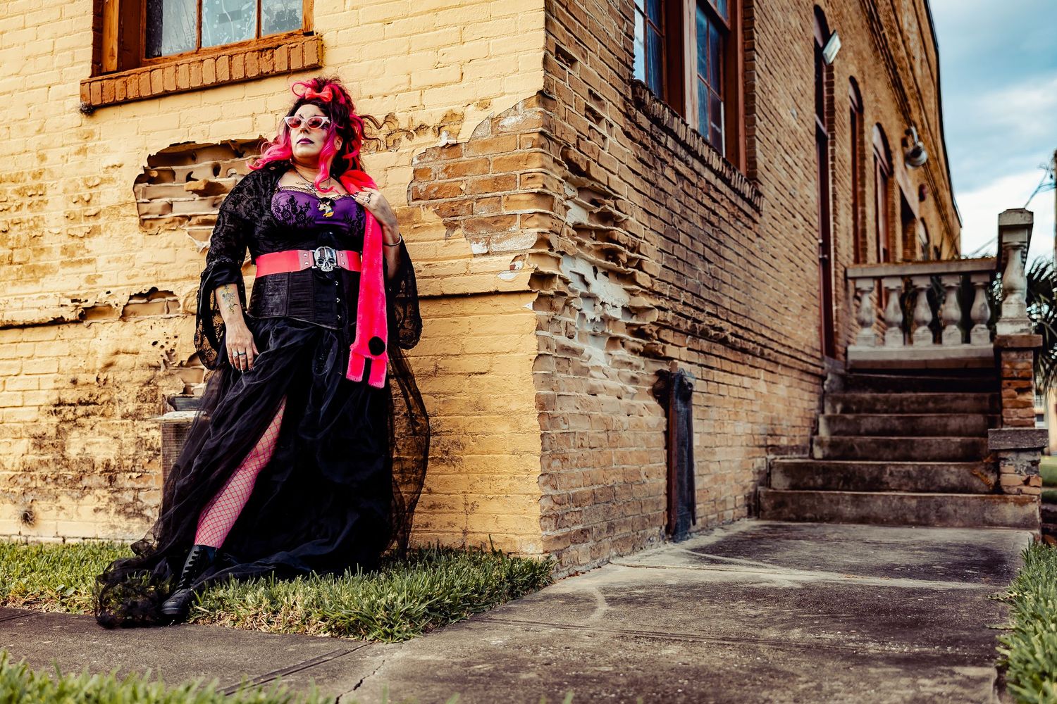 Plus-size woman in dramatic black gothic gown with red accents poses against a rustic brick wall.