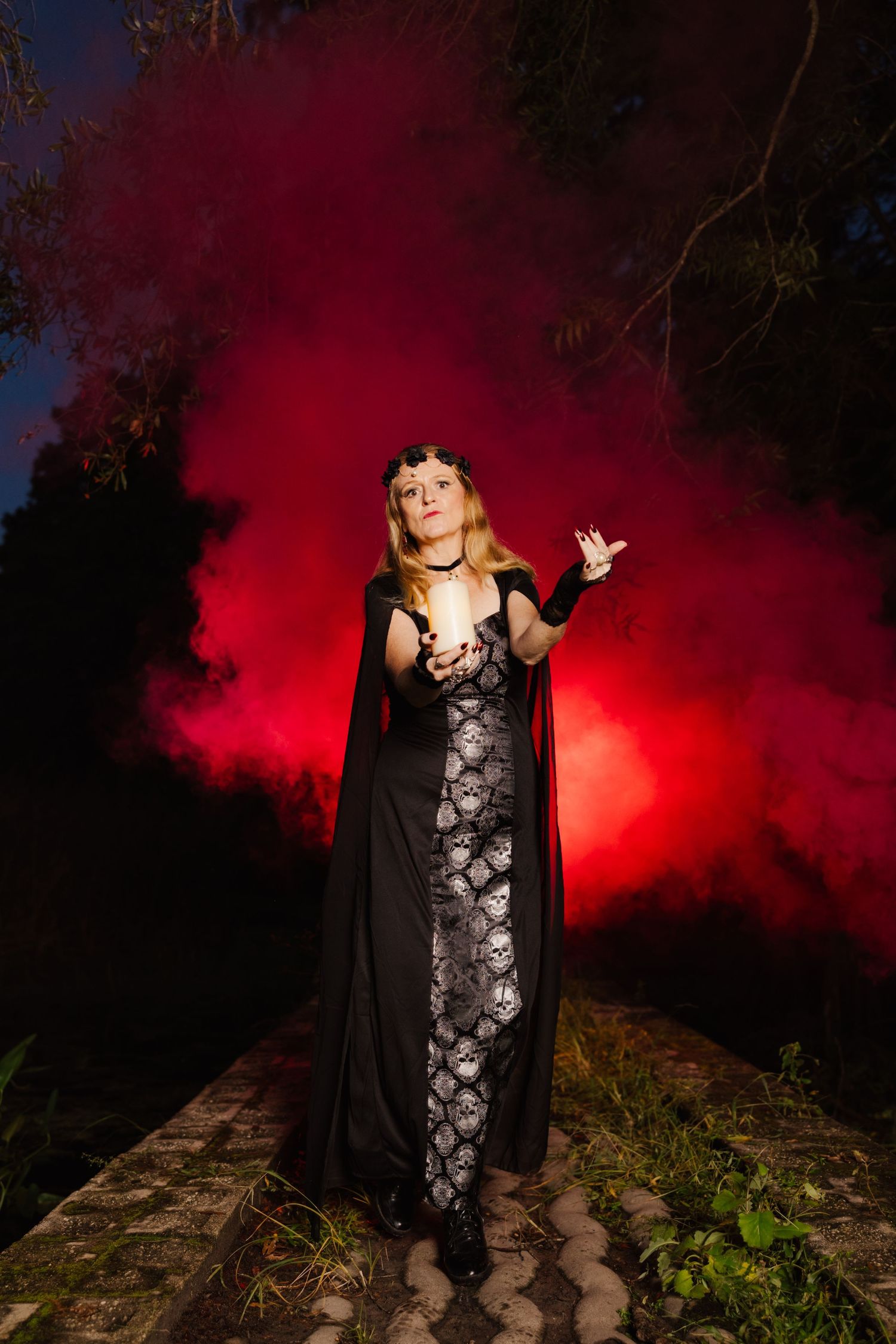 Woman in skull-print dress and black cape poses with glowing orb amid vibrant red smoke in a dark garden.