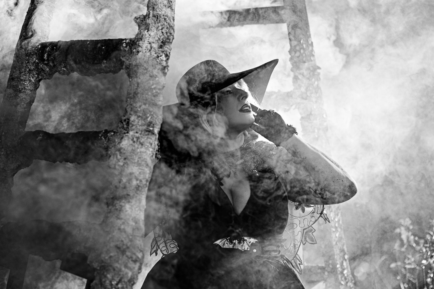 Black and white artistic photo of woman in witch hat surrounded by dramatic billowing smoke.