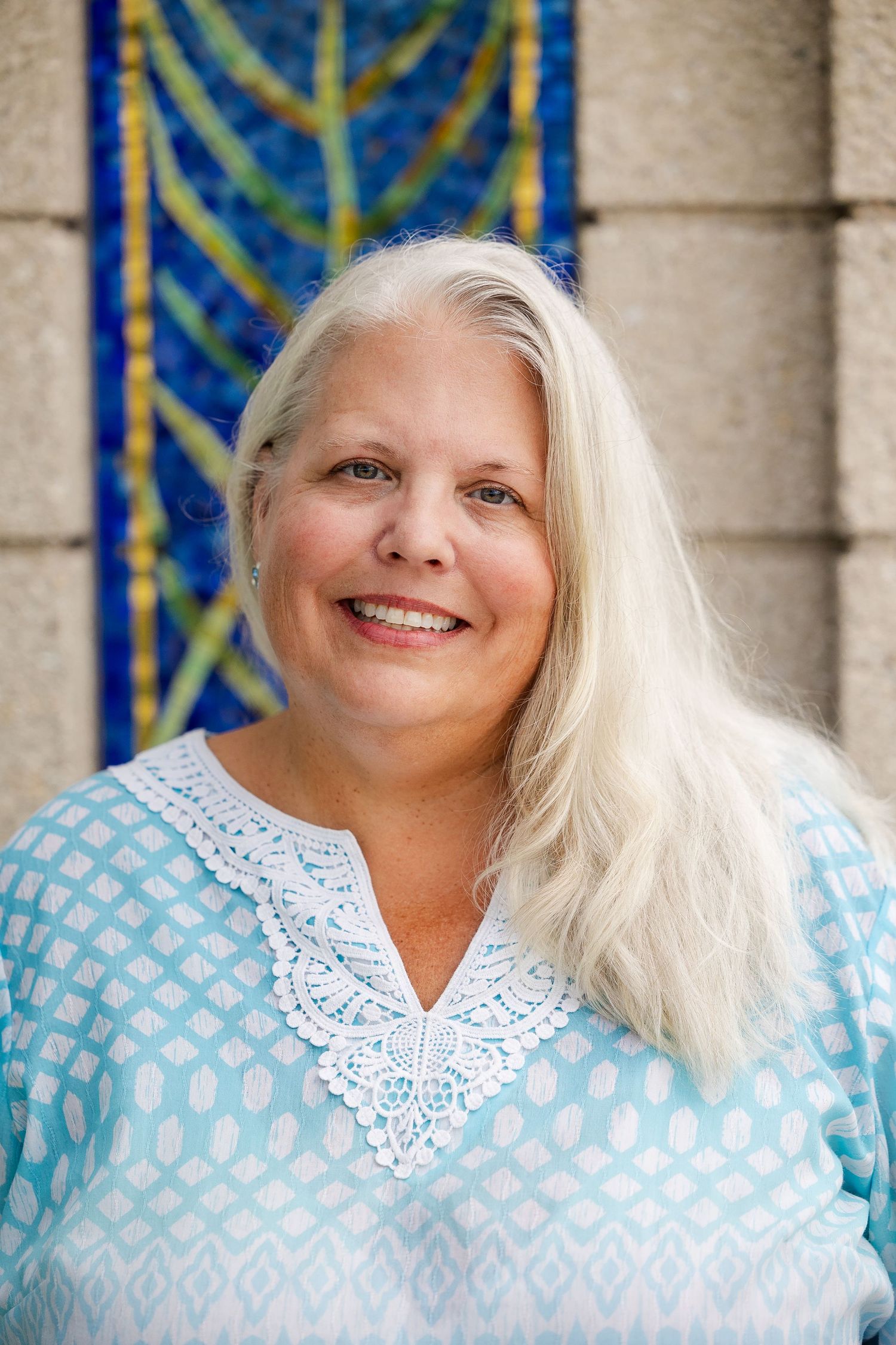 Smiling middle-aged woman with long blonde hair wearing a light blue tunic top, standing in front of a colorful mosaic.