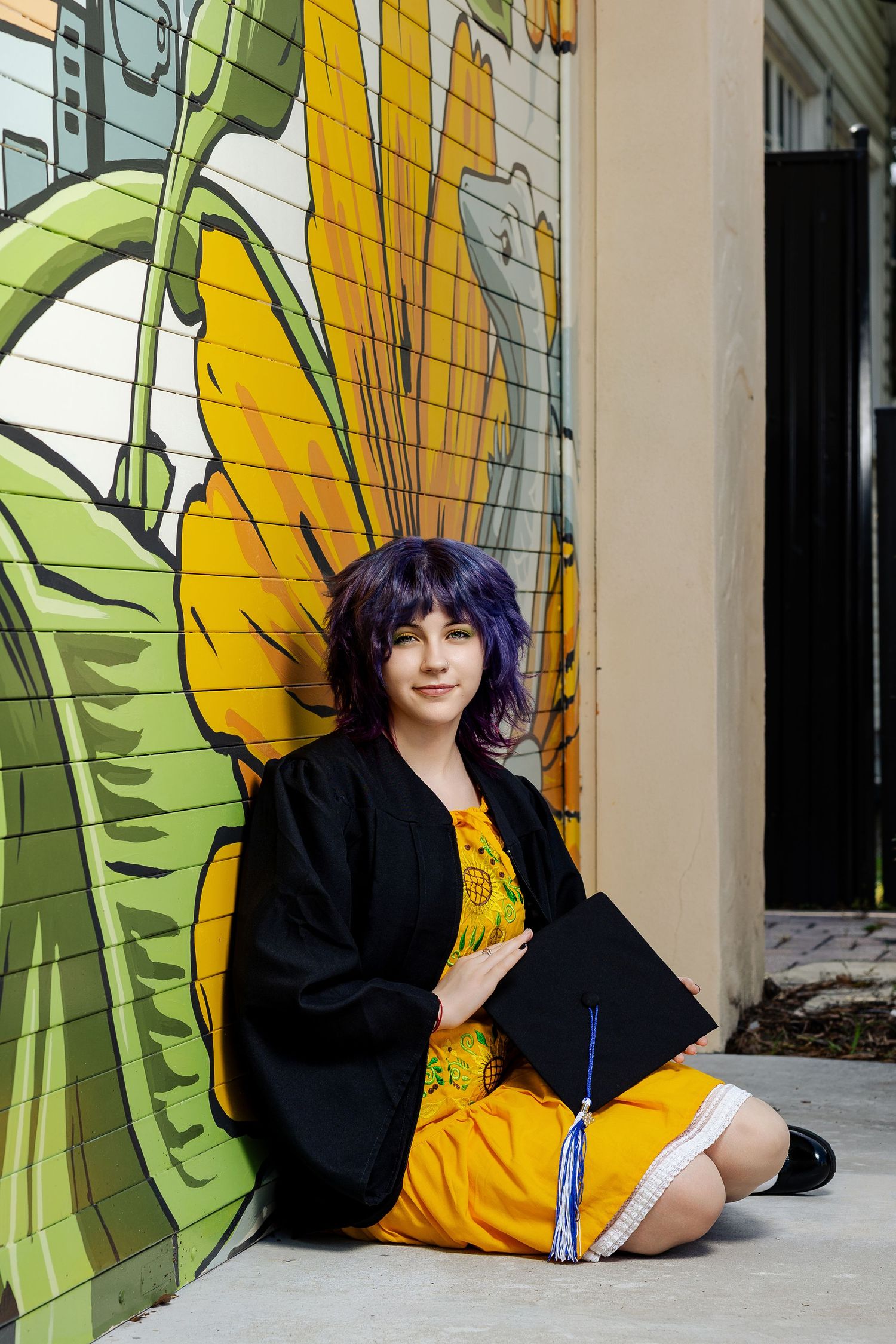 Graduate in black gown and yellow dress holds cap, sitting against colorful sunflower mural wall.