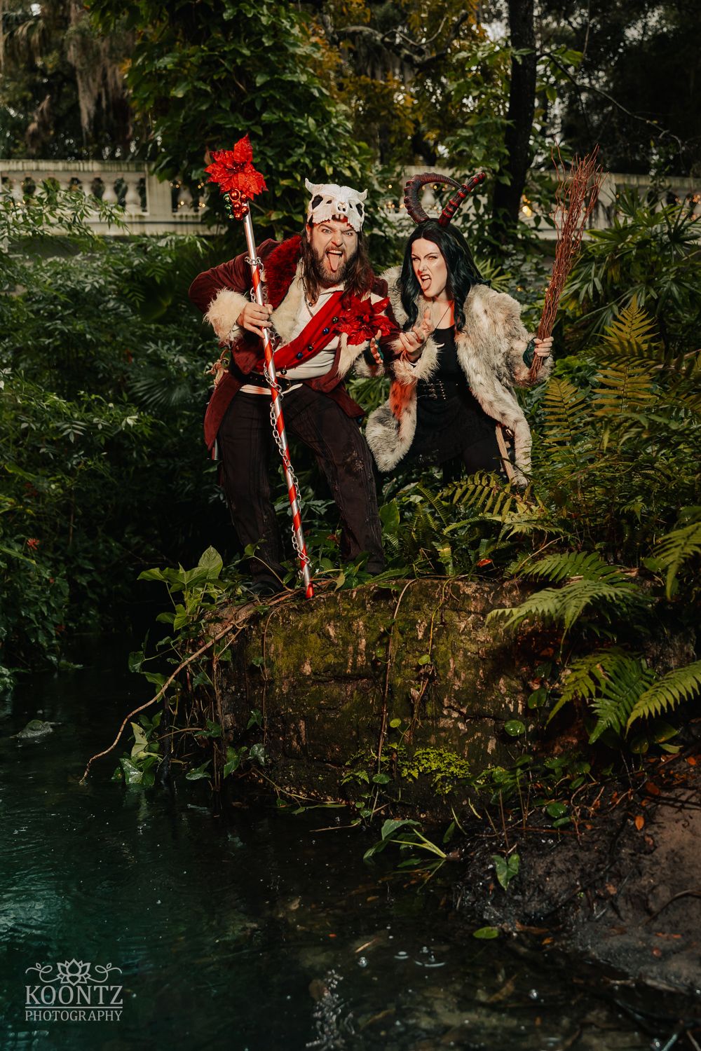 Two cosplayers in dark fantasy costumes leap dramatically over a pond in a lush garden setting.