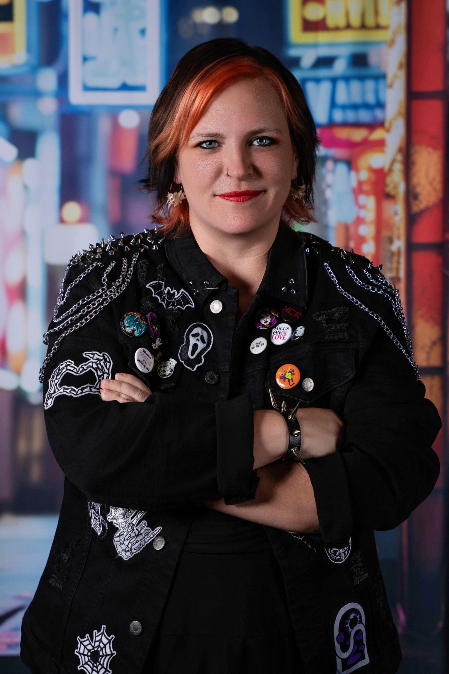 Woman in studded black jacket with pins and red hair poses confidently with arms crossed against colorful city backdrop.