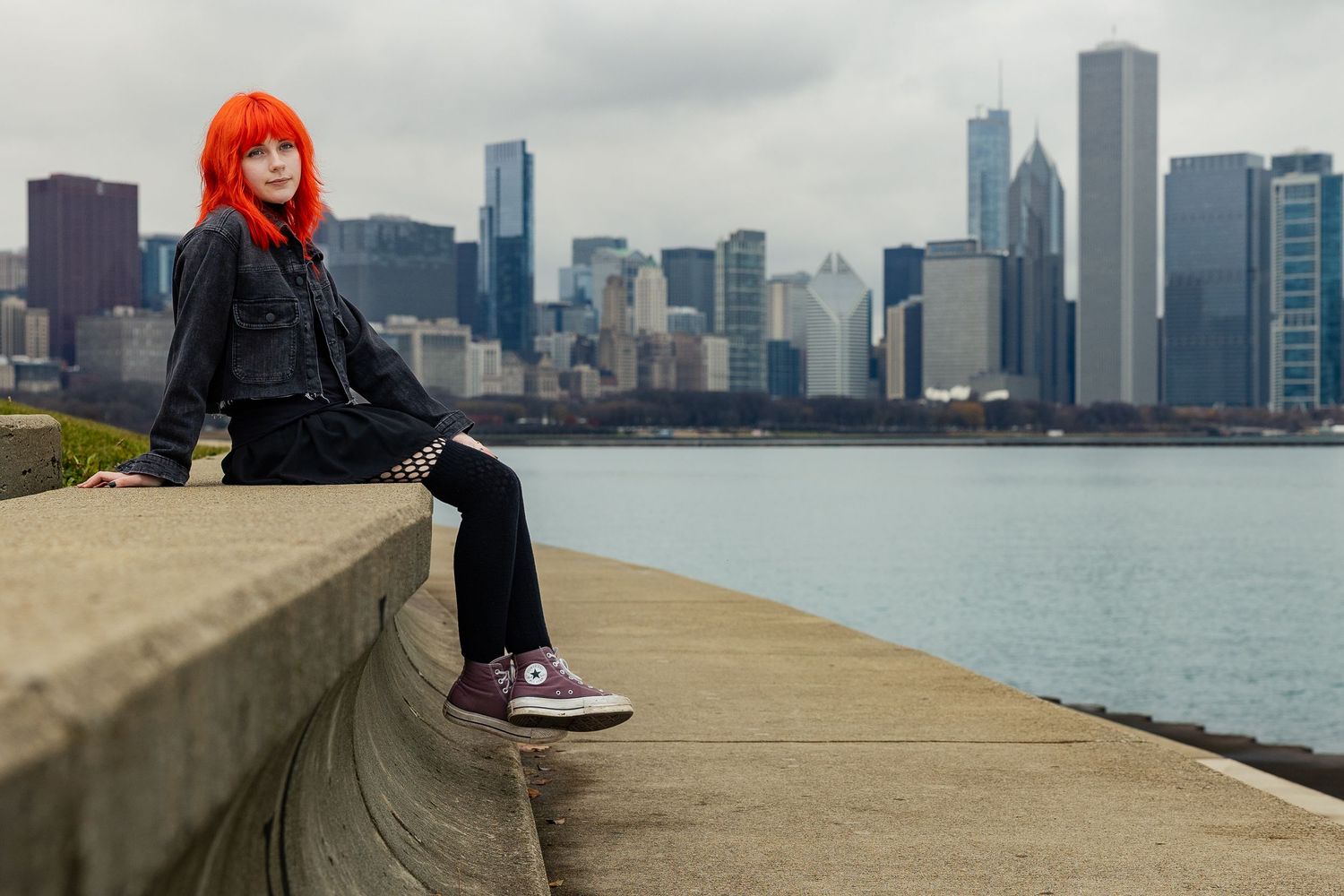 Woman with bright red hair sits on a concrete wall by Chicago skyline waterfront wearing black jacket and purple Converse.