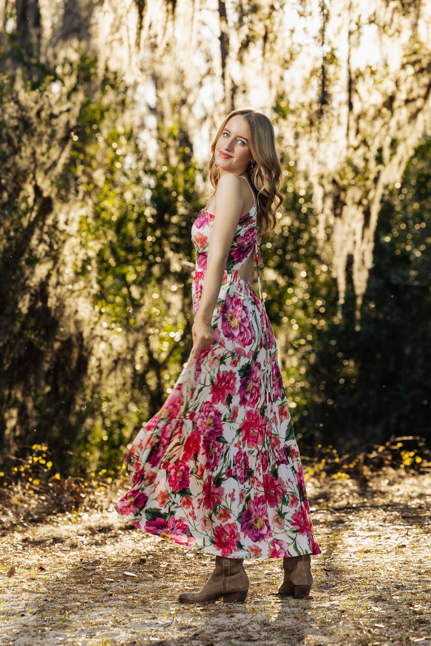 Young woman in floral maxi dress and boots posing outdoors among moss-draped trees in golden sunlight.
