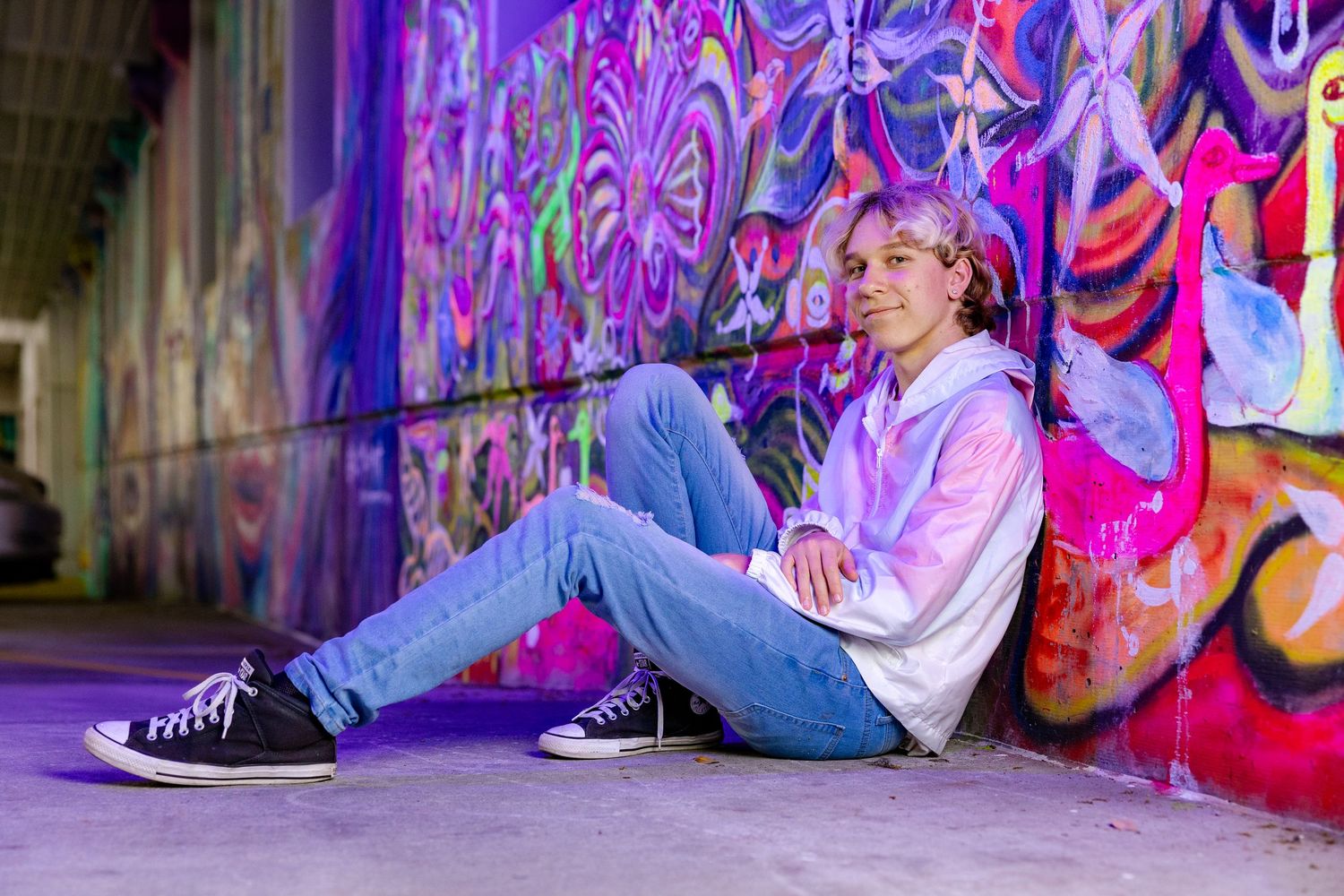 Teen boy sitting against vibrant graffiti wall wearing tie-dye hoodie, jeans, and black Converse sneakers.