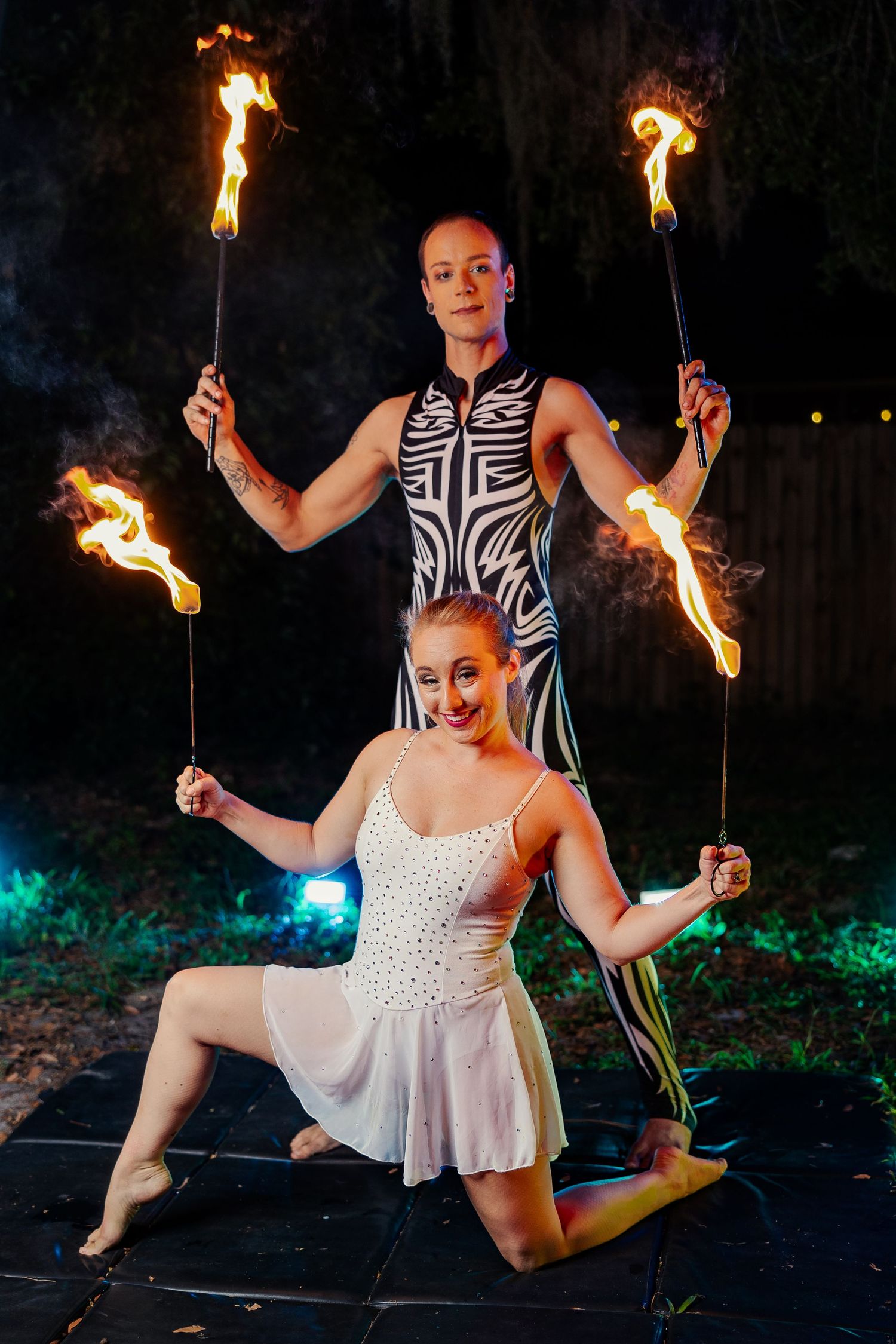 Two fire performers smile at night show, woman kneeling in white dress while partner holds multiple flaming torches.