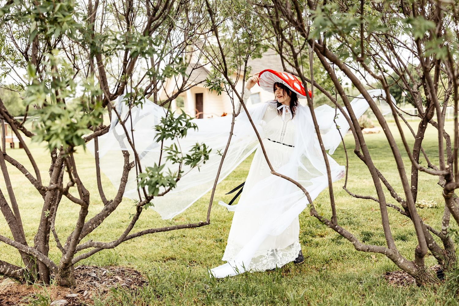 Woman in white dress and red hat poses gracefully among bare trees in a lush green garden.