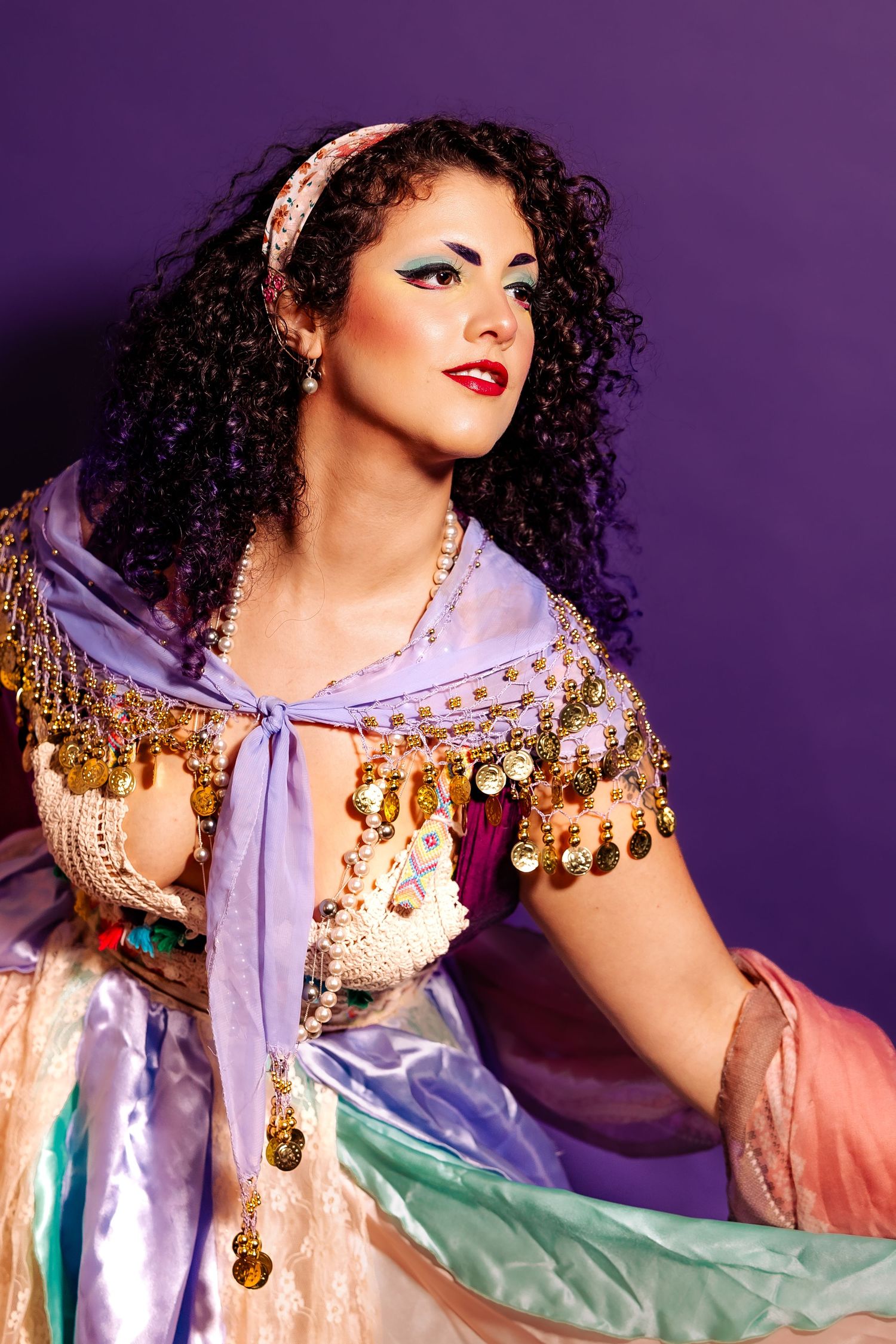 Woman in colorful belly dance costume with gold coins and curly hair poses on purple background.
