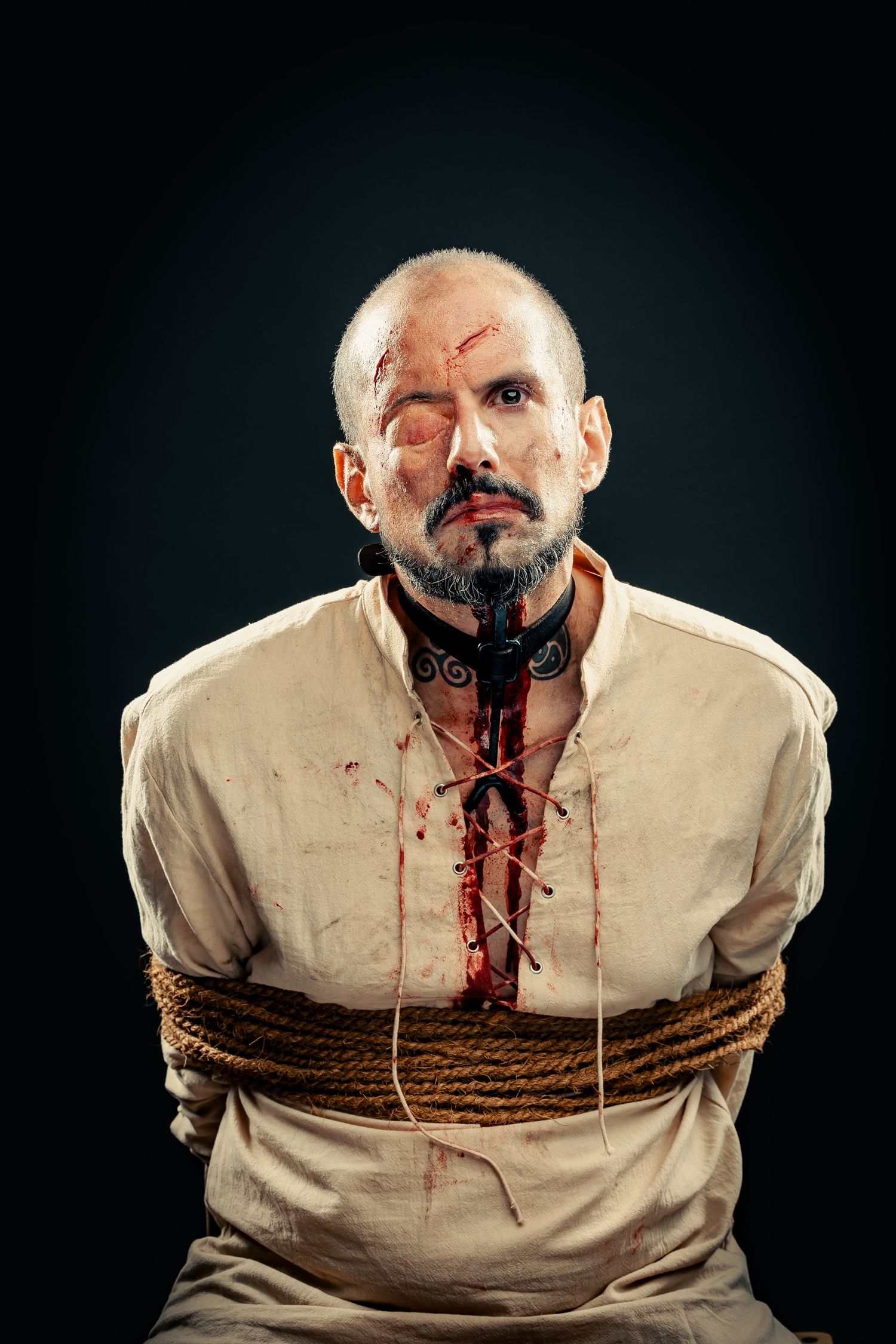 Bald man with bloody wounds and rope bindings portrays a captive prisoner in dramatic costume makeup.